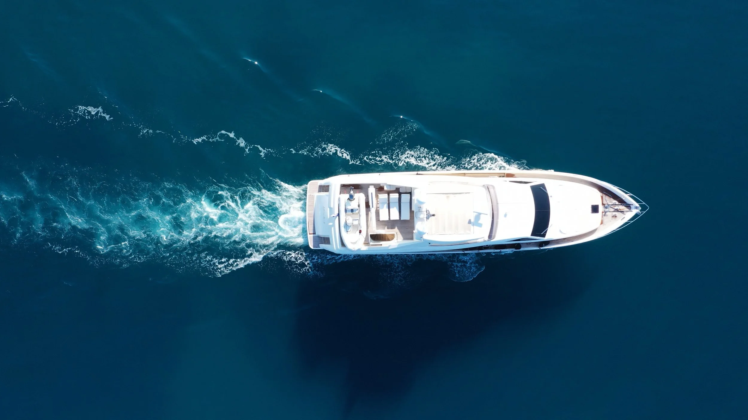 Aerial view of a white yacht sailing on deep blue water, leaving a trail of white foam behind.