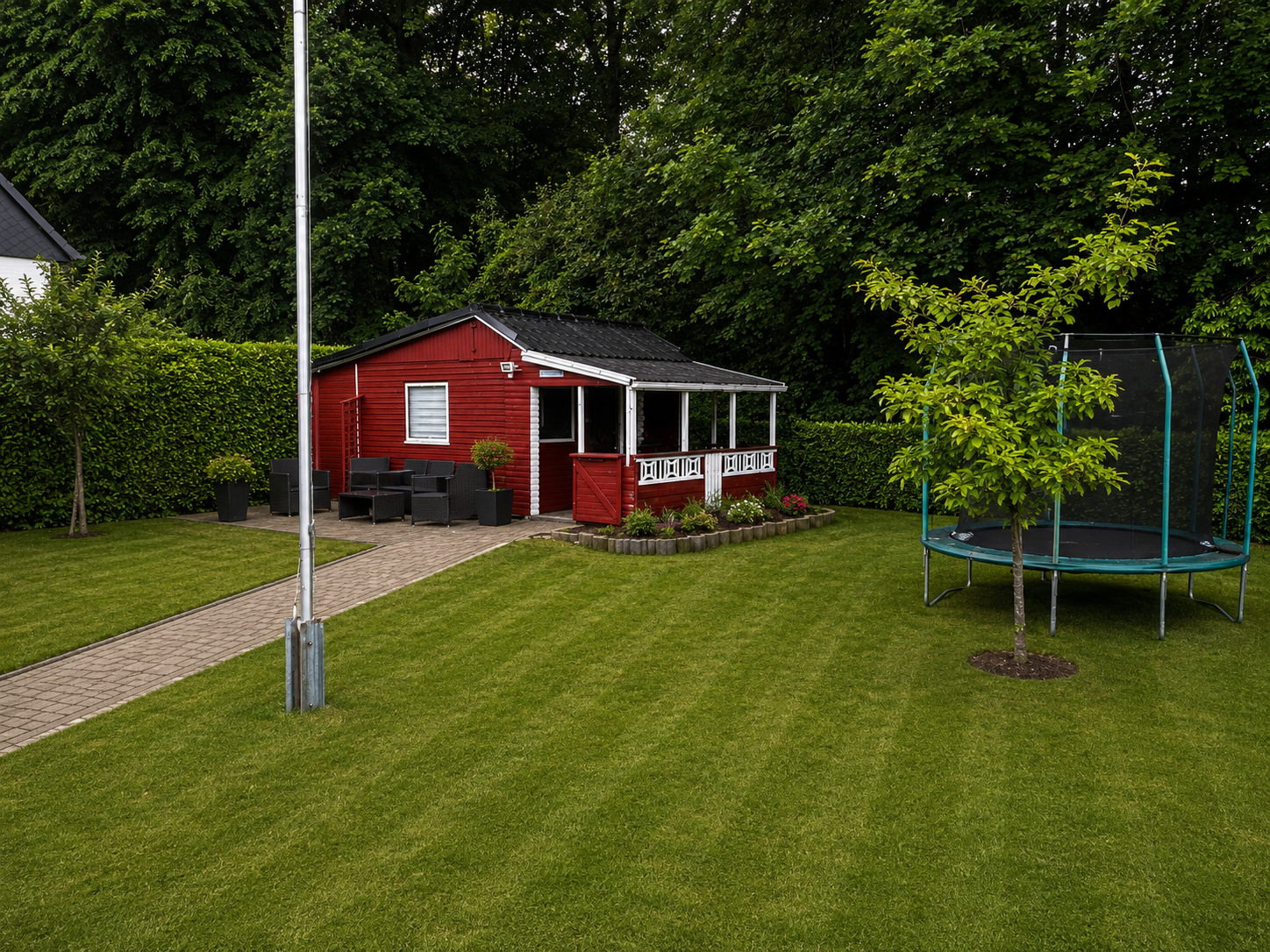 Ein grün gepflegter Garten mit einem roten Gartenhaus, einer sitzgruppe, einem kleinen Baum, einer Hecke, einem Trampolin und einem gepflasterten Weg.