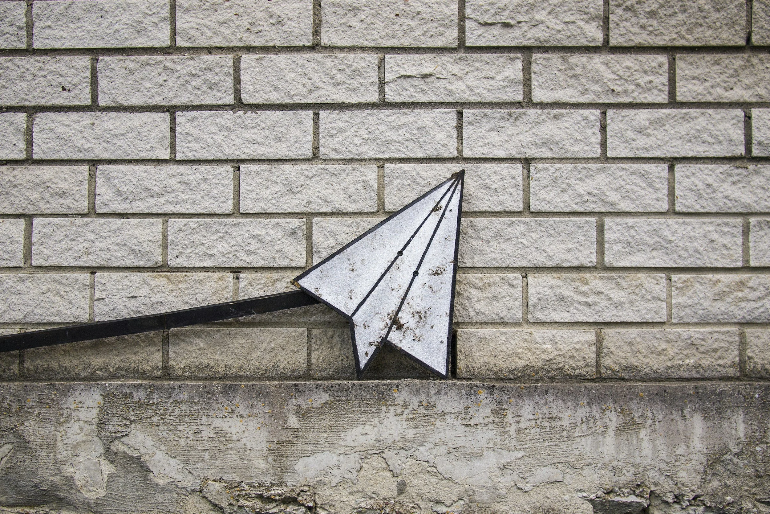 A weathered, white, arrow-shaped street sign attached to a dark metal pole, mounted on a beige brick wall with rough, textured surface and visible mortar lines.