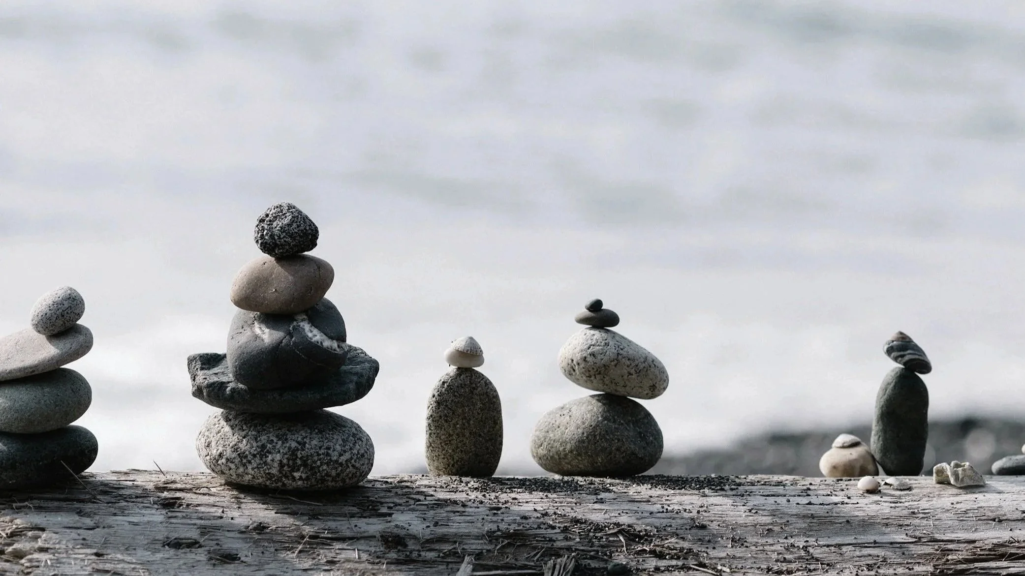 Several stacks of smooth stones of various shapes and sizes balanced on a wooden surface near water, with a cloudy sky in the background.