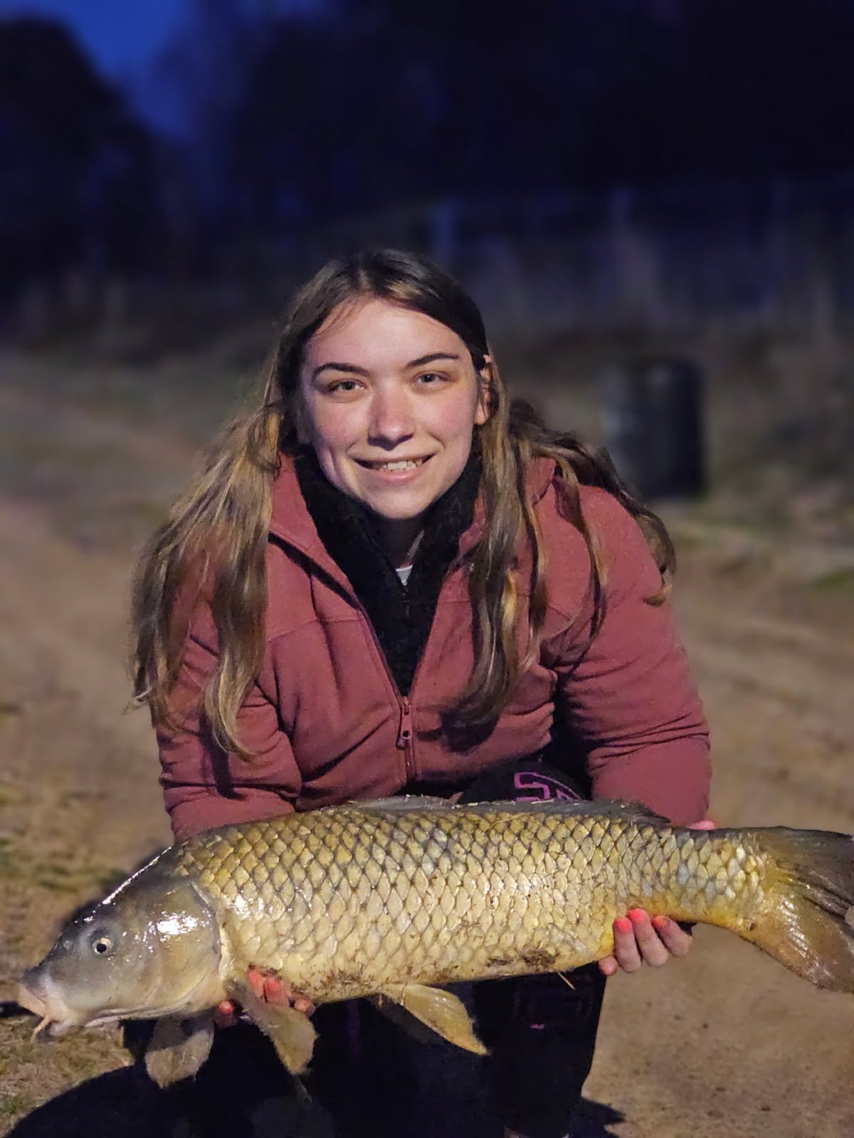 A young woman with long light brown hair smiling, holding a large fish with yellowish scales outdoors at night.