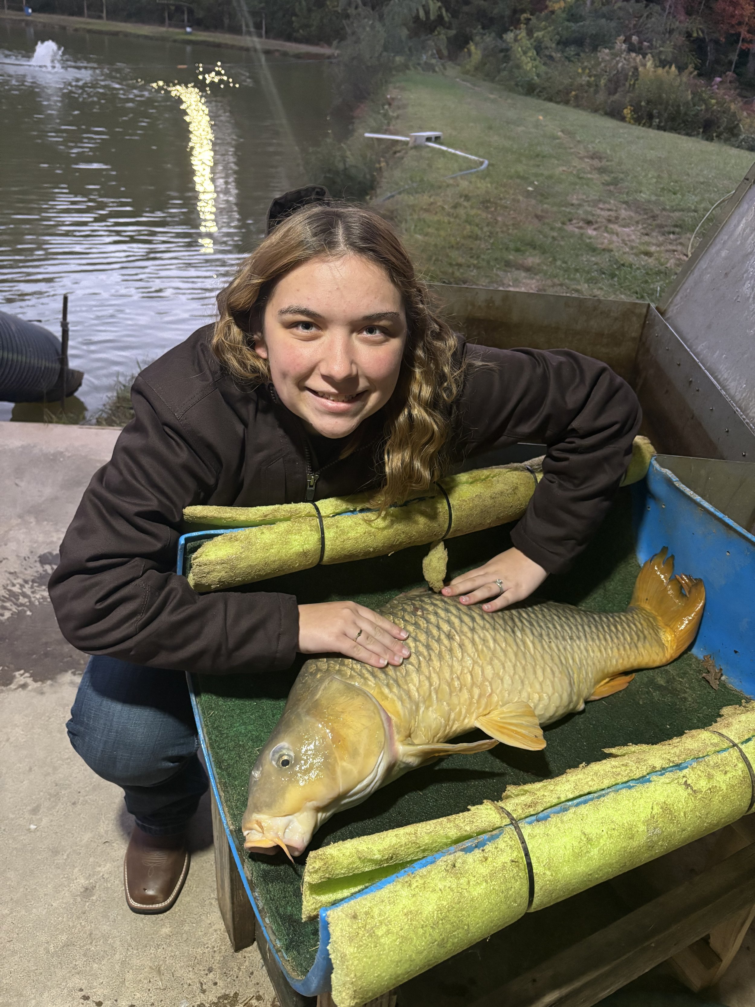 Young woman smiling and posing with a large fish, possibly a carp, on a green padded surface outdoors near a body of water at dusk. The background shows a grassy area, trees, and a fountain on the water.