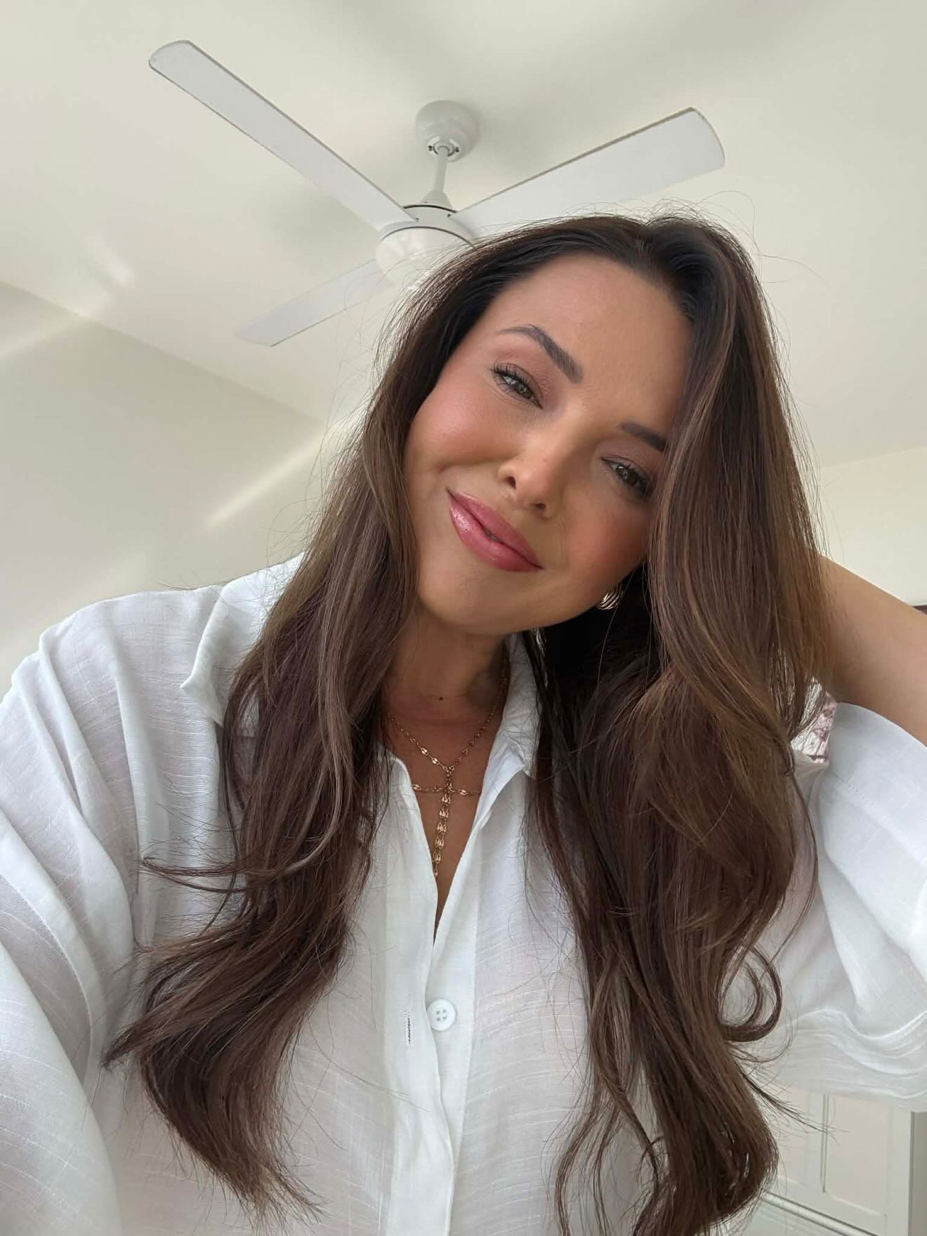 A woman with long, wavy brown hair, wearing a white button-up shirt and gold jewelry, smiling, in a room with a ceiling fan.