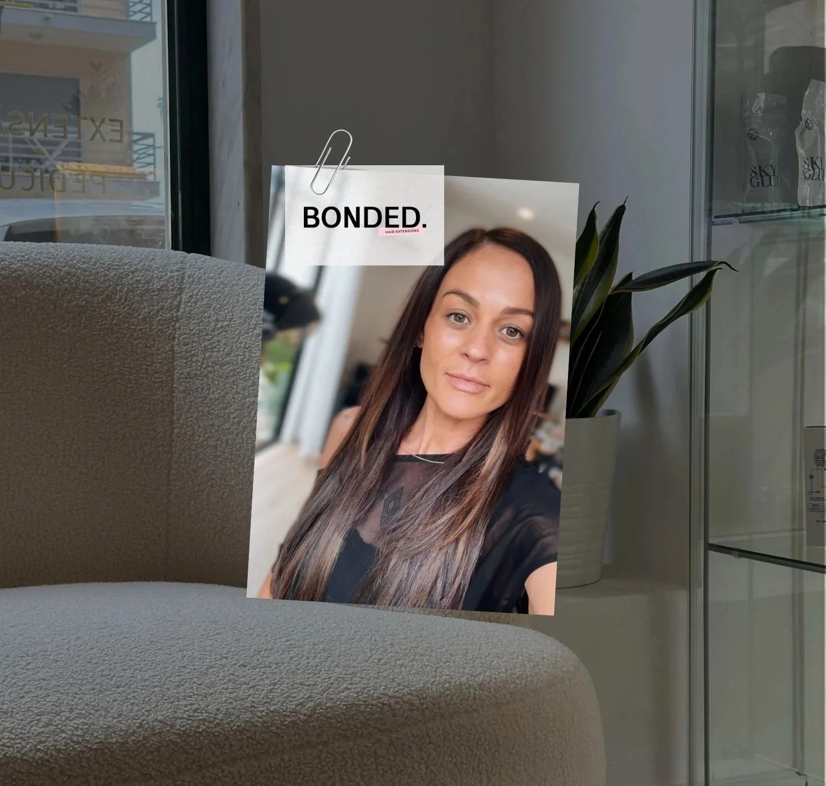 The founder with long brown hair and a black shirt taking a selfie inside a modern building with large glass windows and green plants, with a signboard in the background that reads "BONDED."