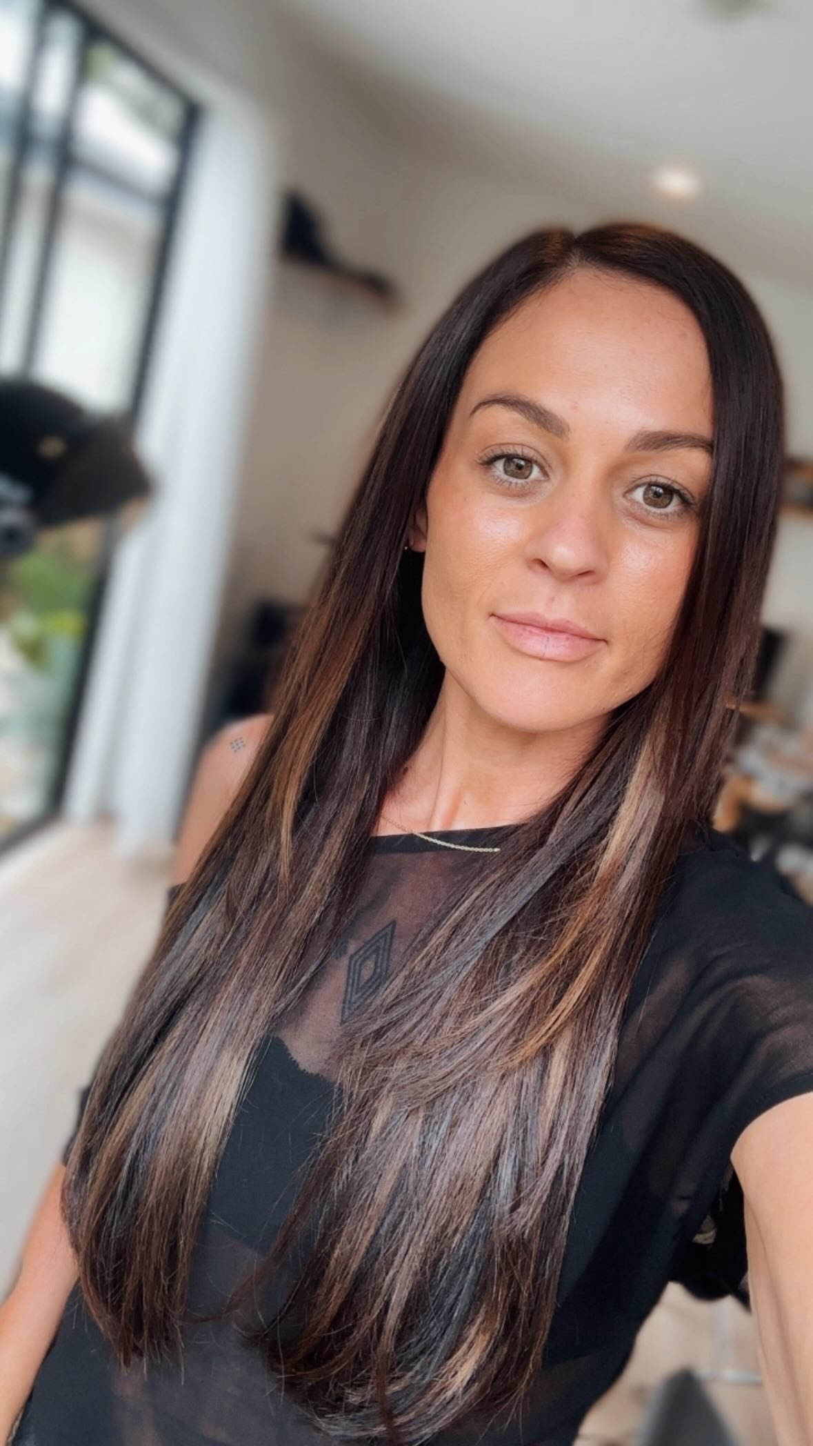 The founder with long, straight  hair taking a selfie in a well-lit room with large windows and a blurred background.