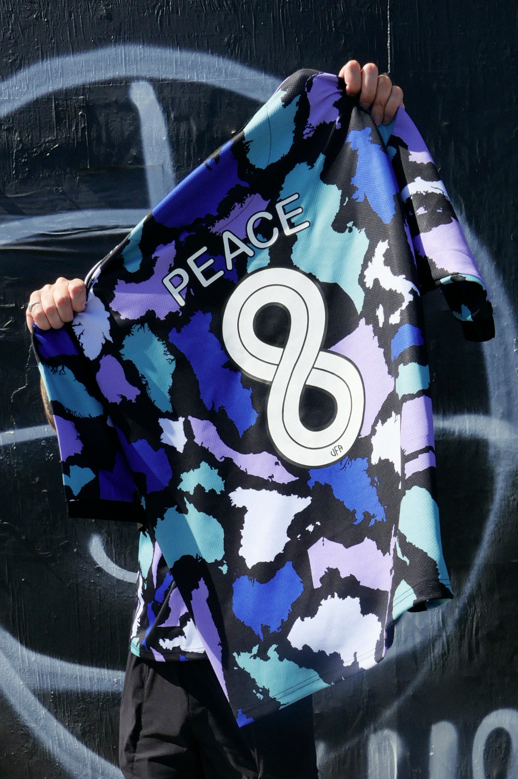 A person holding a colorful sports jersey in front of a black wall with graffiti, featuring the words "PEACE" and the number 96 with a stylized infinity symbol.