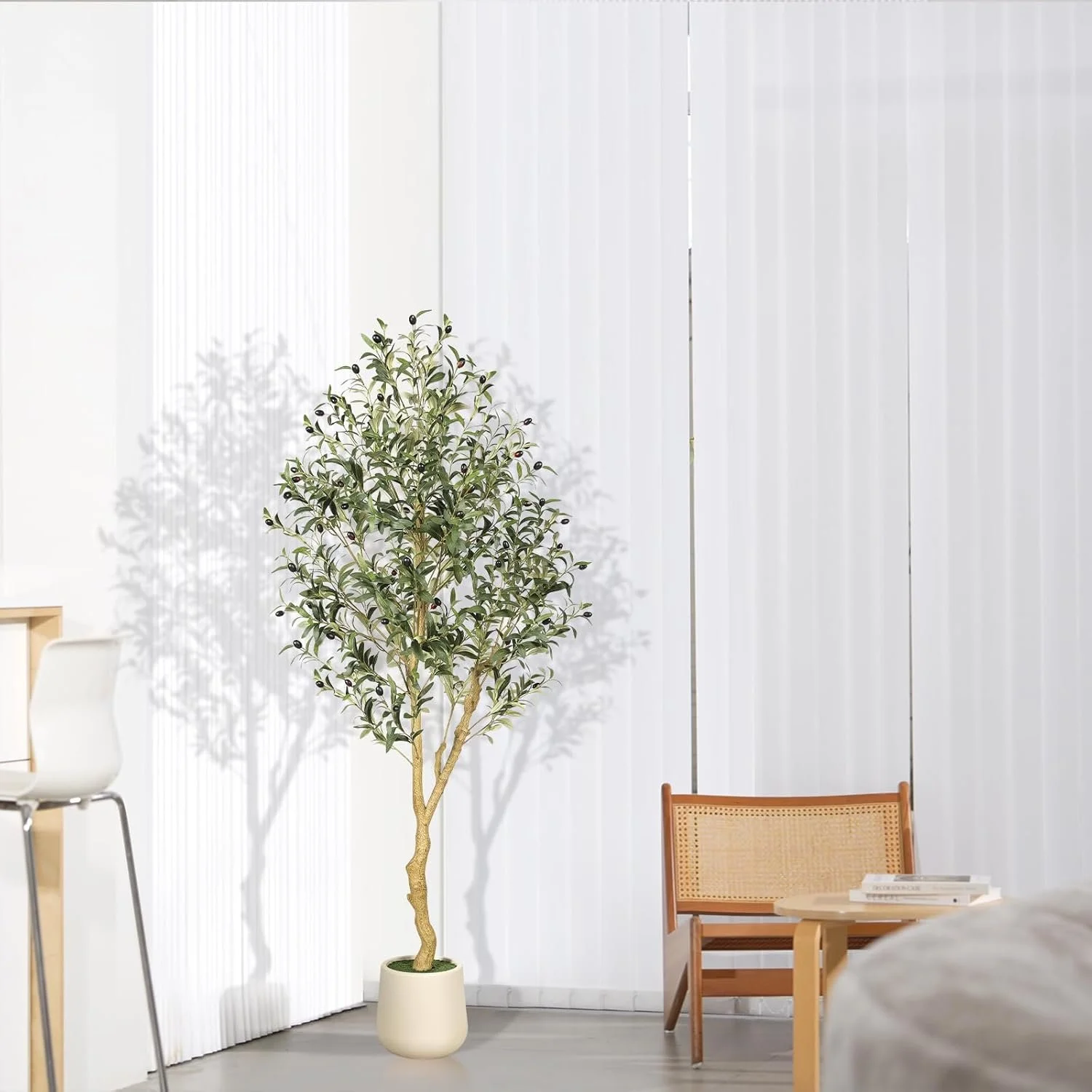 An indoor space with a tall potted olive tree, a white chair, a wooden bench with books, and vertical white window blinds.