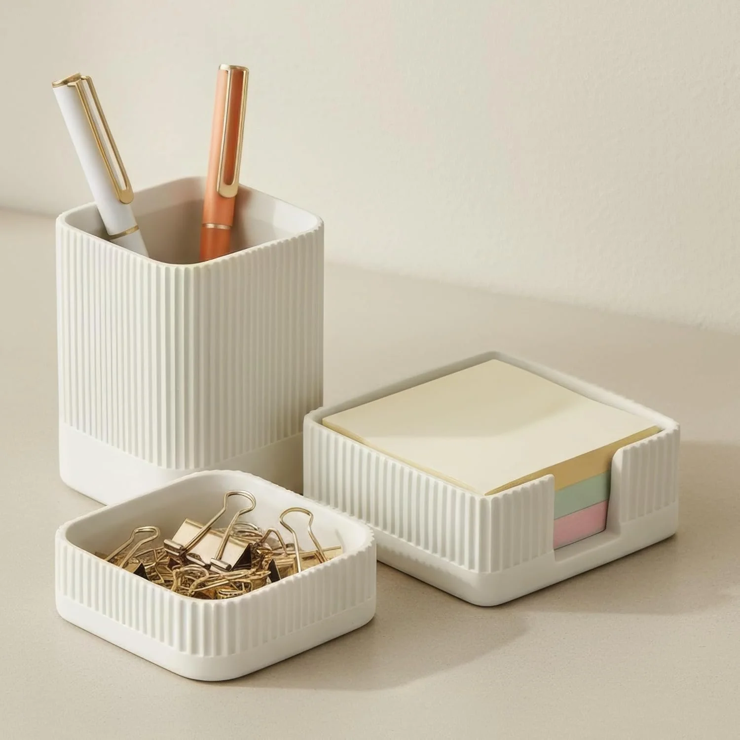 White desk organizers containing pens, paper clips, sticky notes, and paper binders on a beige surface.