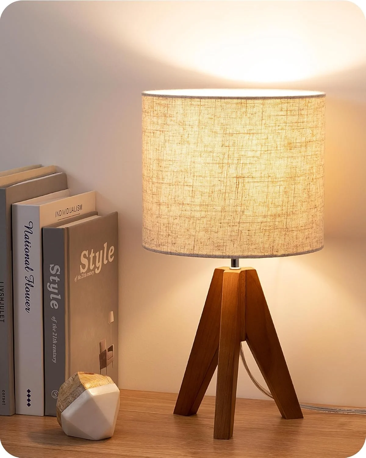 A table lamp with a beige fabric shade and a wooden tripod base, placed on a wooden surface next to a few books and a decorative geometric object, with a soft warm light.