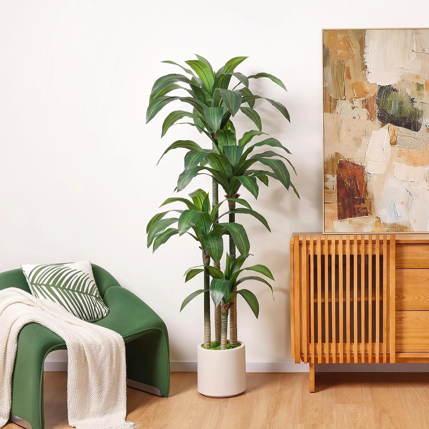 A tall potted plant with green elongated leaves in a white pot on a wooden floor. To the left, part of a green chair and a white blanket with a pillow featuring green palm leaves. To the right, a wooden cabinet and an abstract painting.
