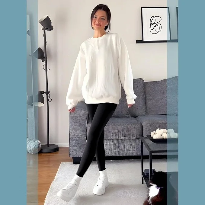 A woman standing in a living room, wearing a white oversized sweatshirt, black leggings, and white sneakers, with a relaxed pose and a slight smile.