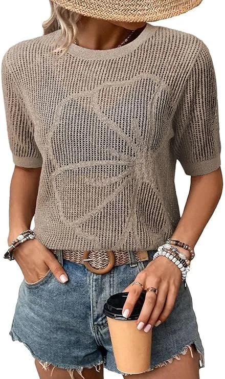 A woman wearing a beige knit top with a large butterfly design, denim shorts, a brown belt, a wide-brim straw hat, and multiple bracelets. She is holding a coffee cup.