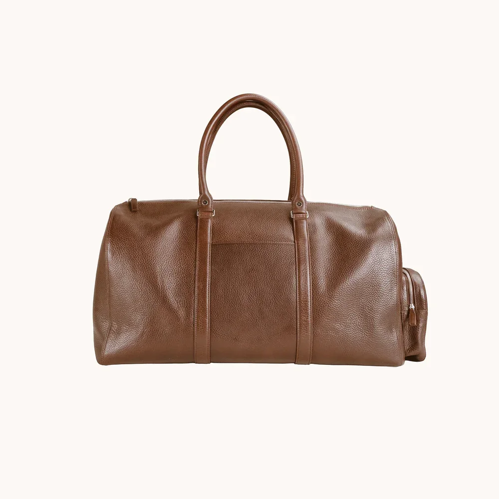 A brown leather duffel bag with two handles and a side zipper pocket, set against a plain white background.