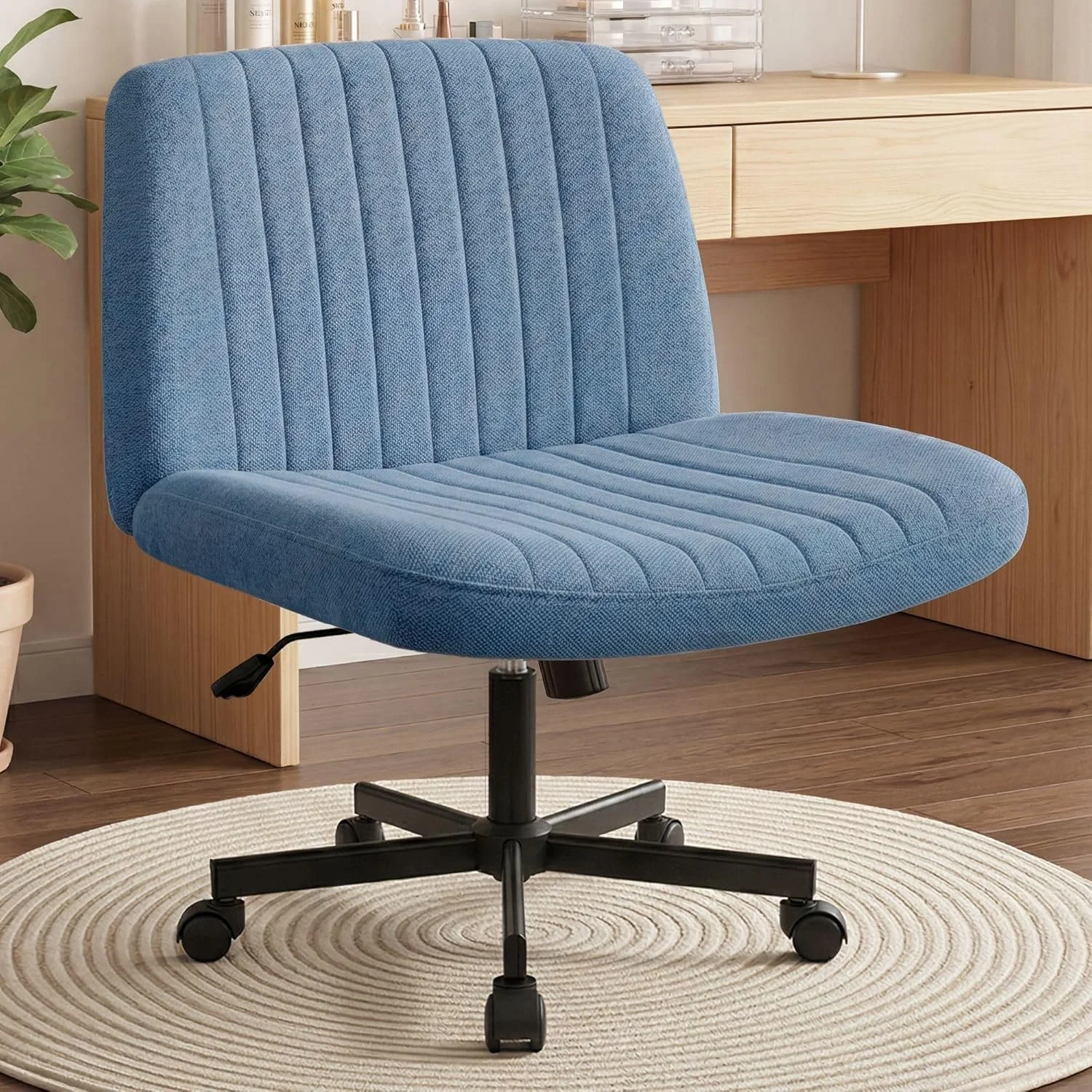 A blue upholstered office chair with a swivel base and caster wheels, placed on a round beige rug in a room with wooden flooring and a wooden desk in the background.