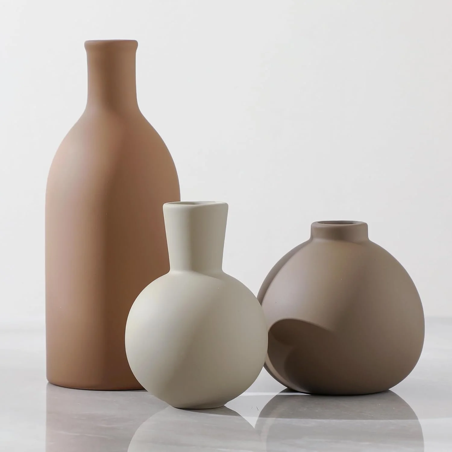 Four ceramic vases in beige, white, and cream colors, with minimalist designs, arranged on a reflective surface against a plain white background.
