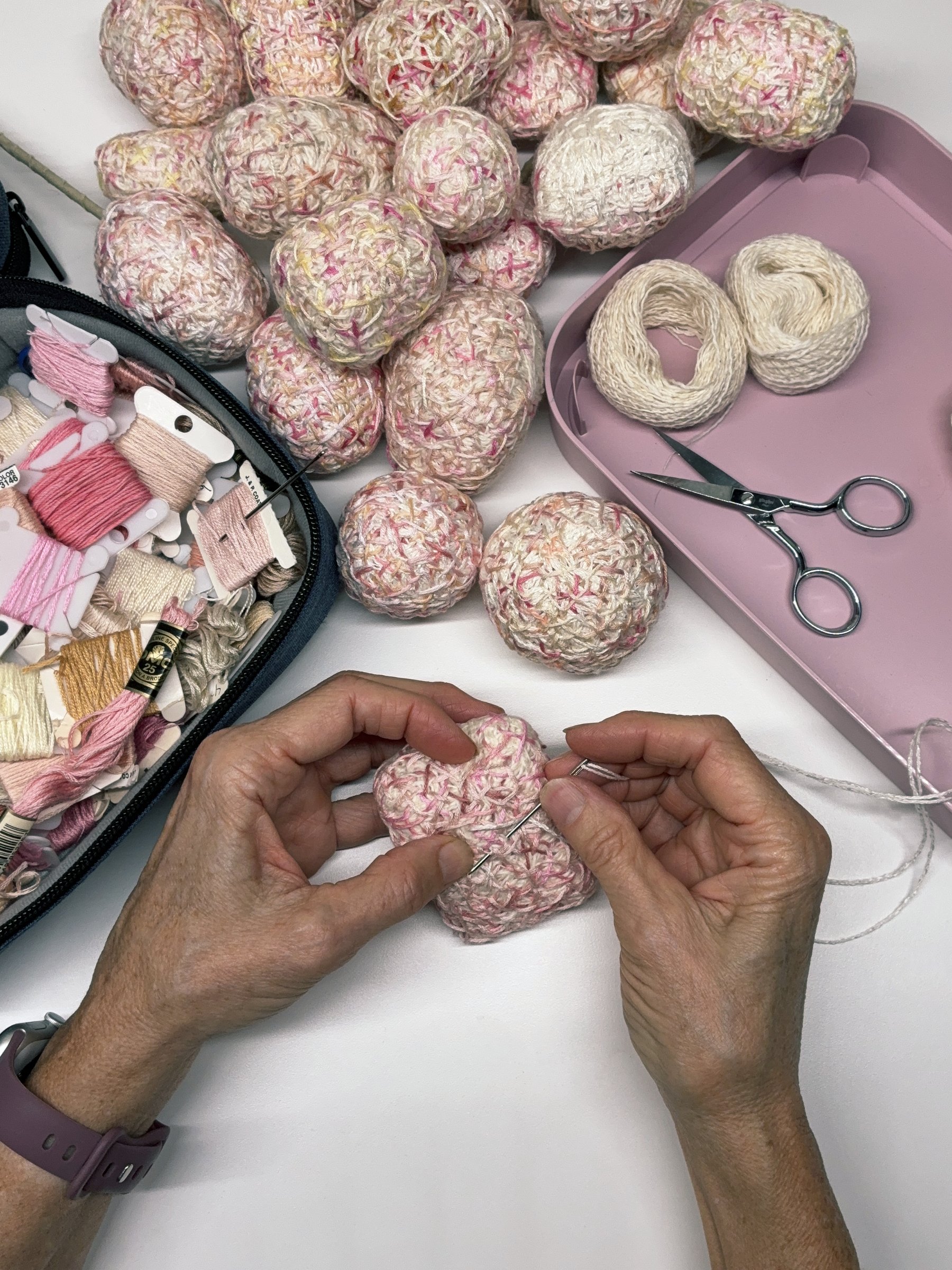 A person embroidering a laceweight-yarn covered item with pink and cream yarns, surrounded completed cocoons, scissors, and embroidery supplies on a white surface.