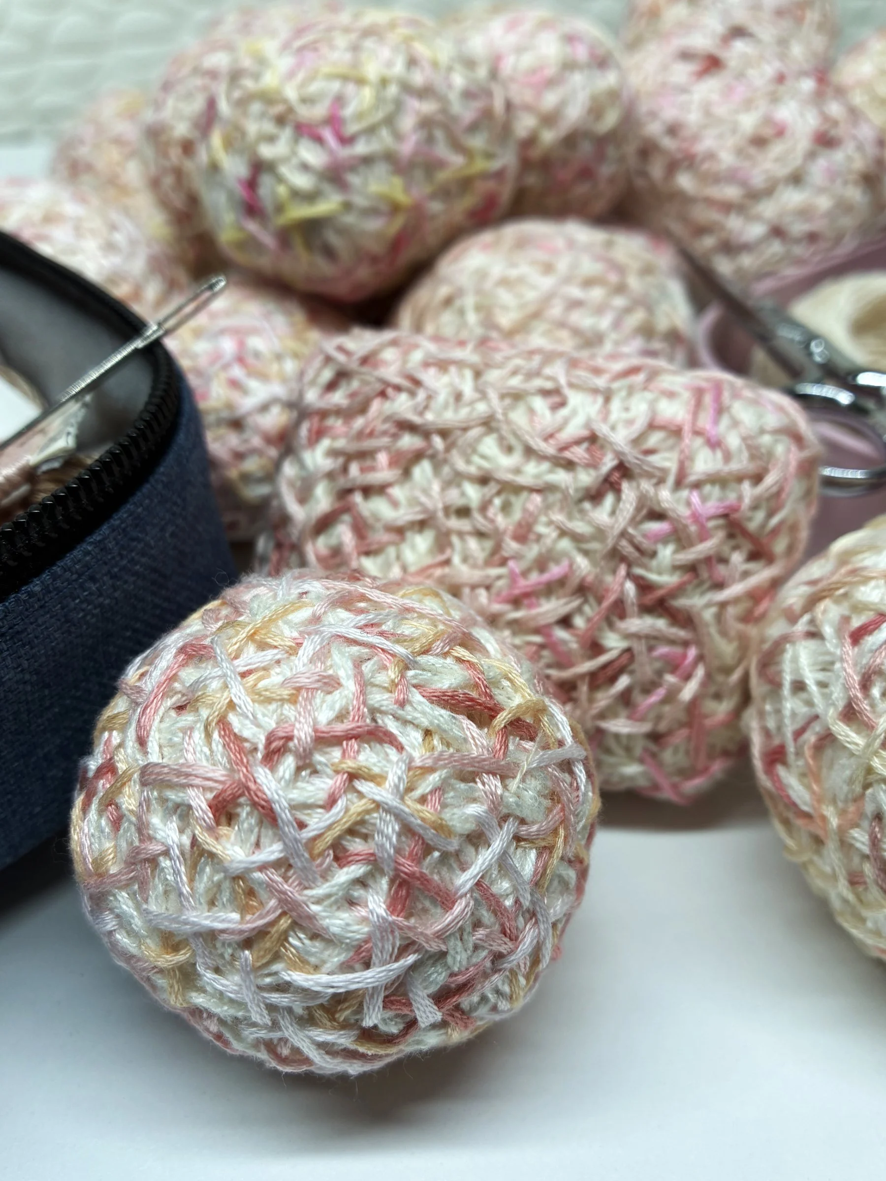 Close-up of pastel-colored yarn balls in pink, yellow, and white, with a sewing needle partially visible at the left and a pair of scissors at the right, on a white surface.