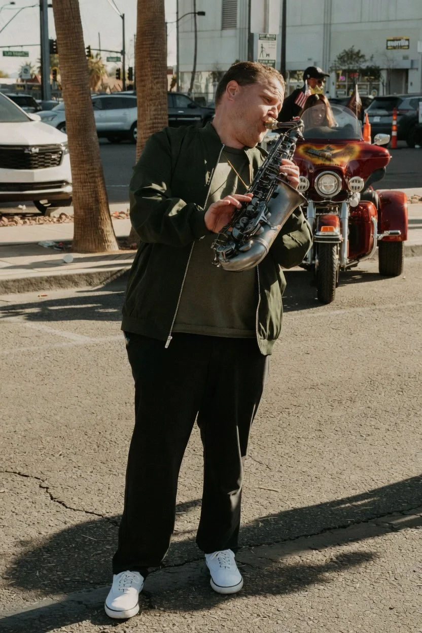 A man playing a saxophone on a city street with parked cars, trees, and a red motorcycle in the background.