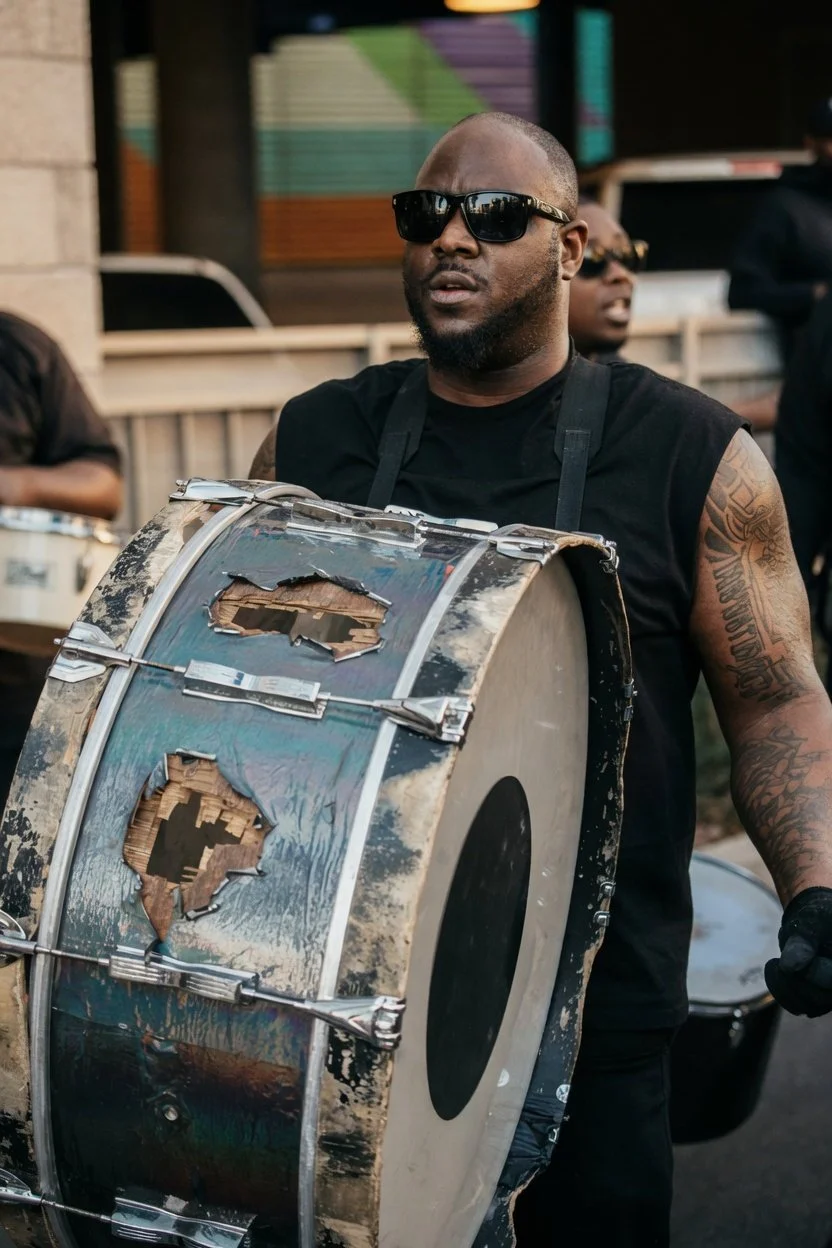 A man wearing sunglasses and a black sleeveless shirt is holding a large, battered drum with holes in its surface. Other musicians are visible in the background.