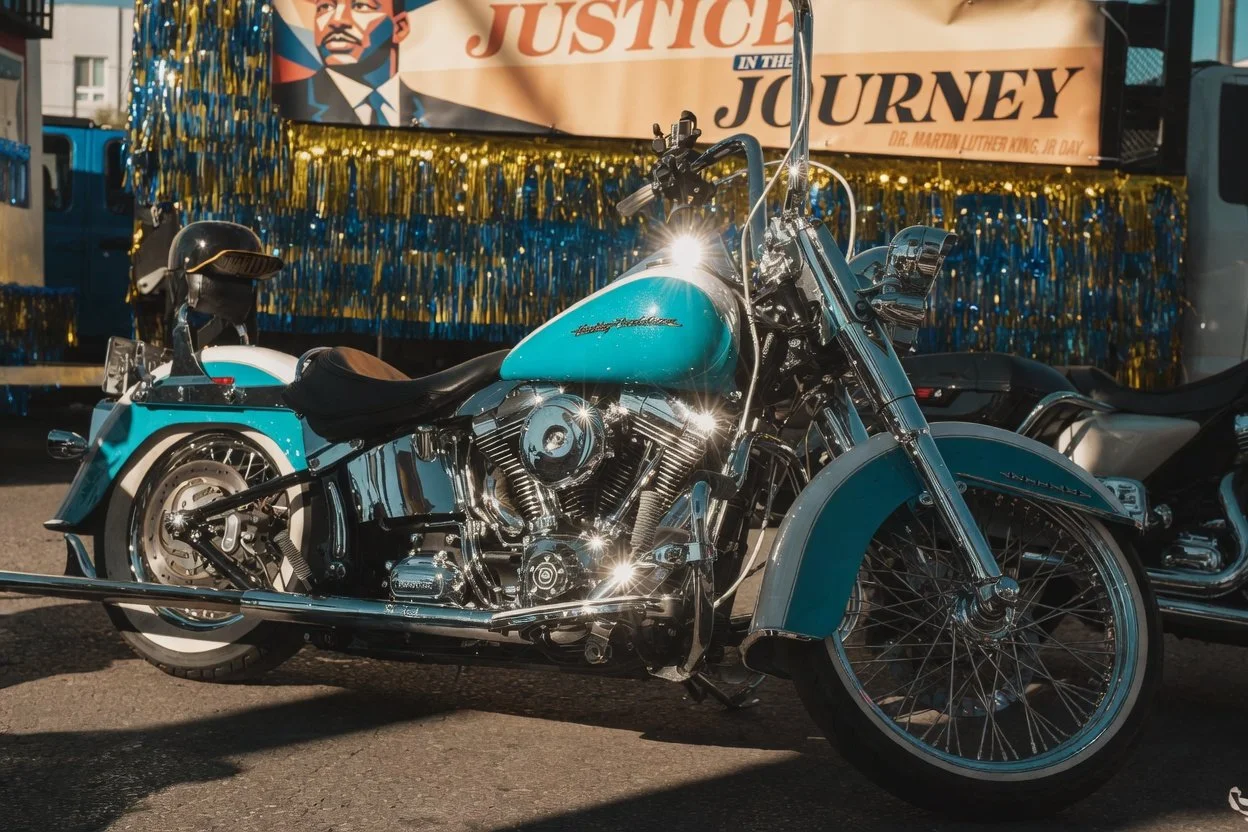 A vintage blue and chrome Harley-Davidson motorcycle parked outdoors during a celebration event with a banner reading "Justice in the Journey" and a picture of Dr. Martin Luther King Jr. in the background.