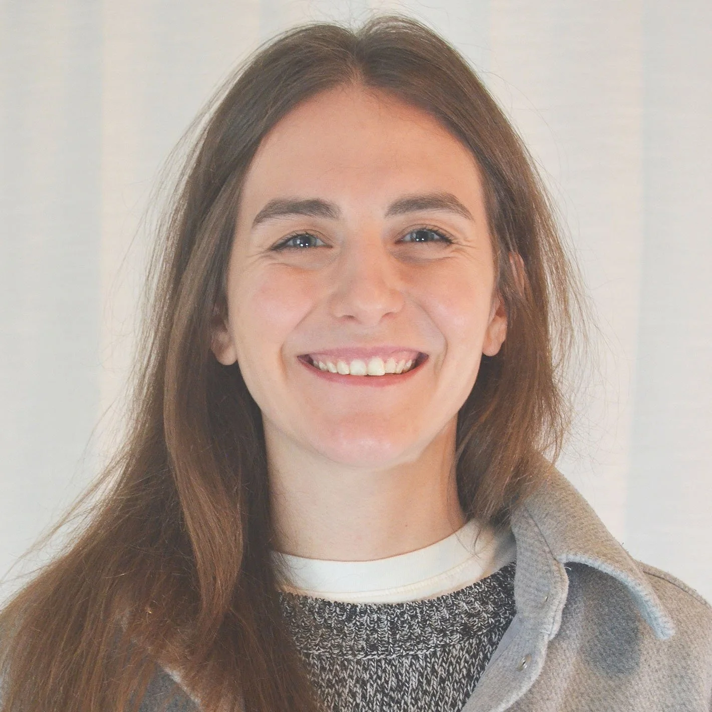A smiling woman with long brown hair, wearing a gray sweater and a beige coat, standing against a plain light-colored background.