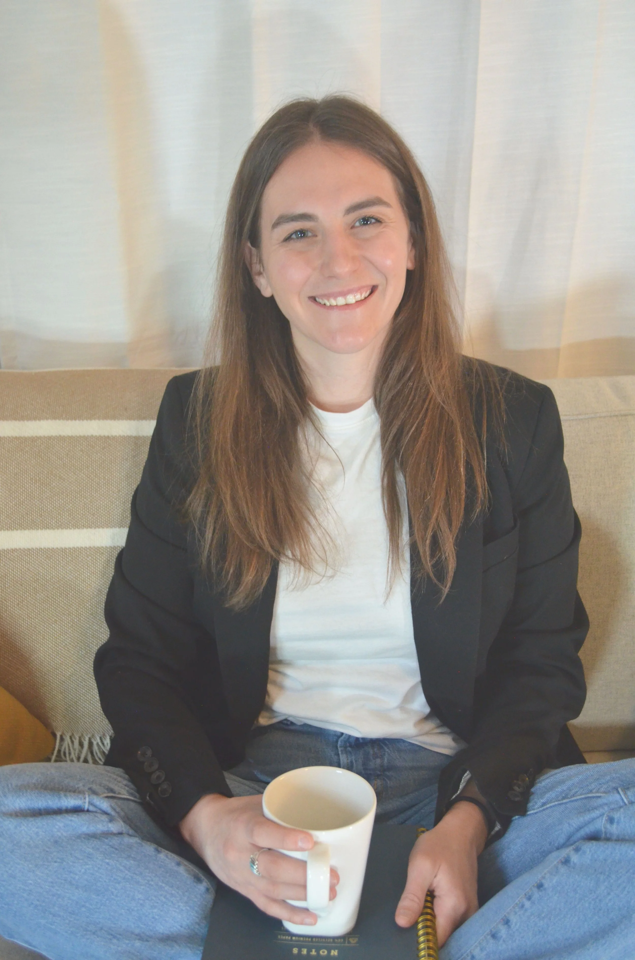 A young woman with long brown hair, wearing a white t-shirt and a black blazer, sitting on a beige couch with white curtains in the background. She is smiling, holding a white mug in her right hand, with a black notebook on her lap, and wearing jeans.