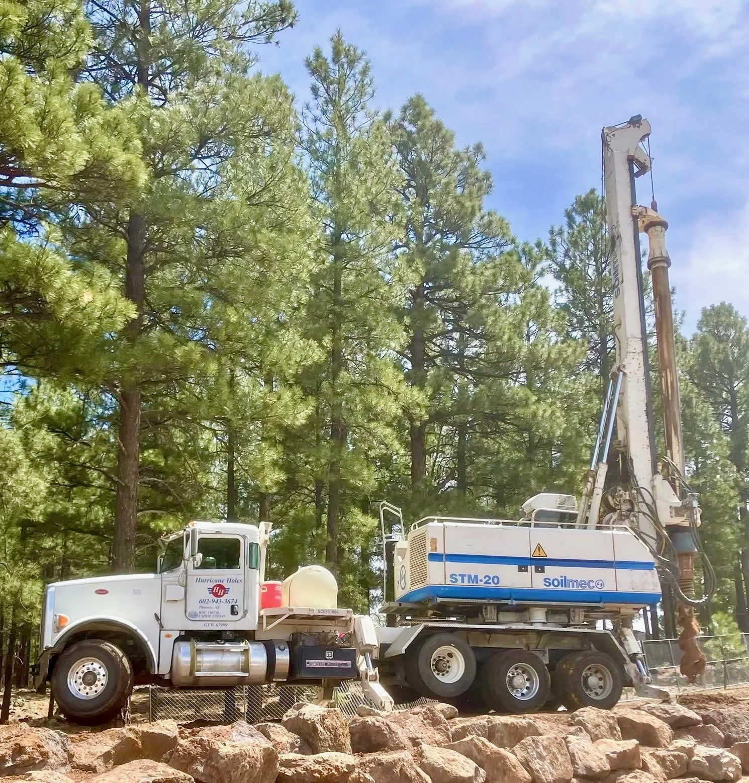 A large drill rig working among tall pine trees in Flagstaff, AZ under a partly cloudy blue sky.