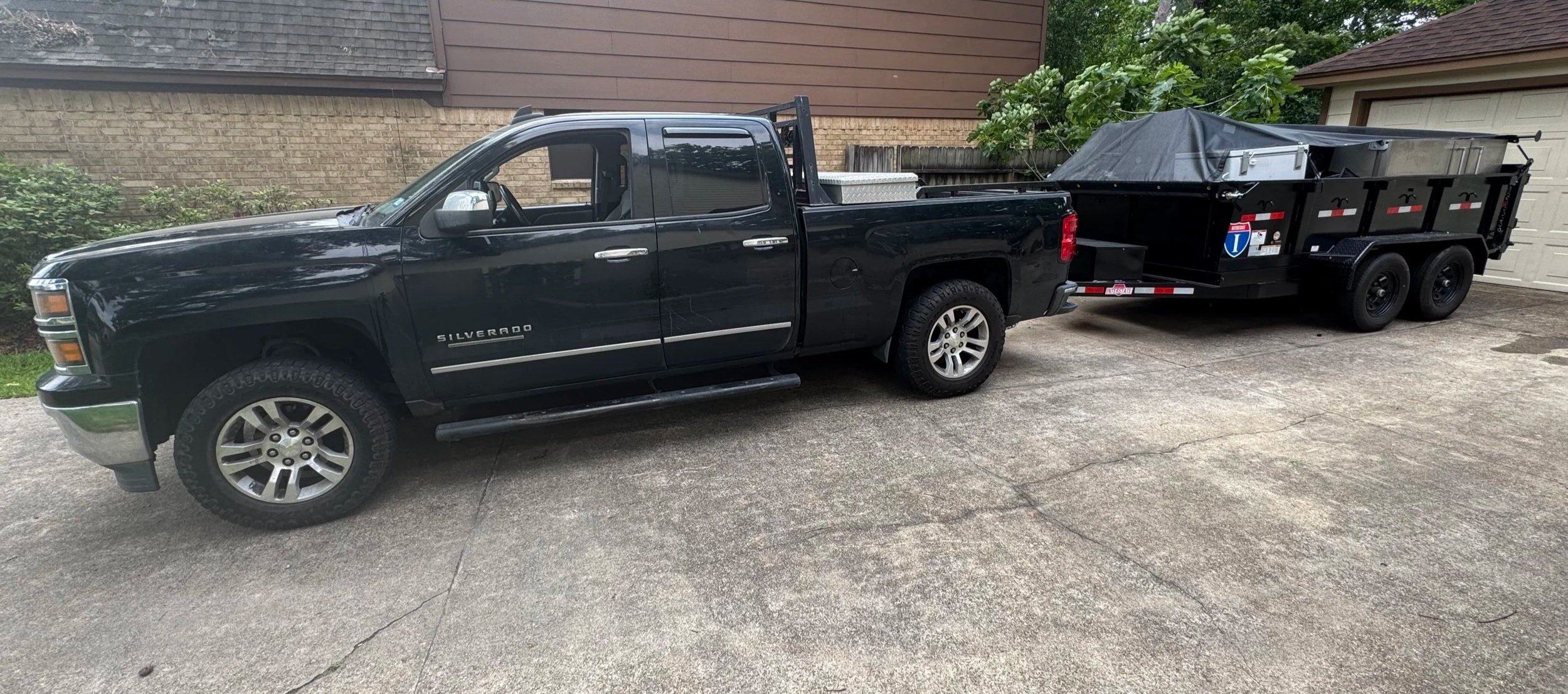 Black Chevrolet Silverado pickup truck with a trailer attached, parked in a driveway next to a house with a brick and wooden exterior.