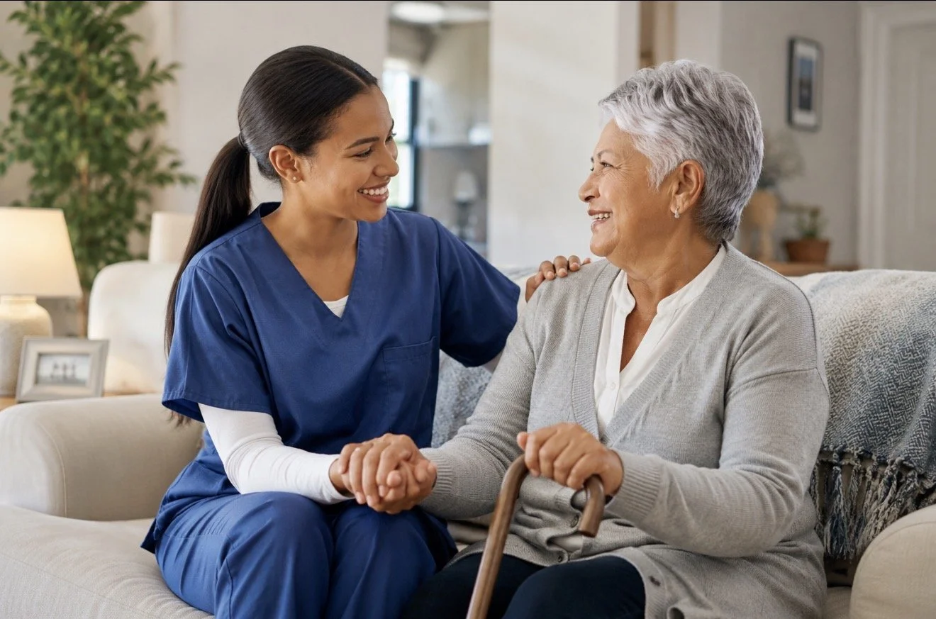 A young female caregiver in blue scrubs smiling and sitting next to an elderly woman with short gray hair, holding a walking cane and smiling at each other in a living room.