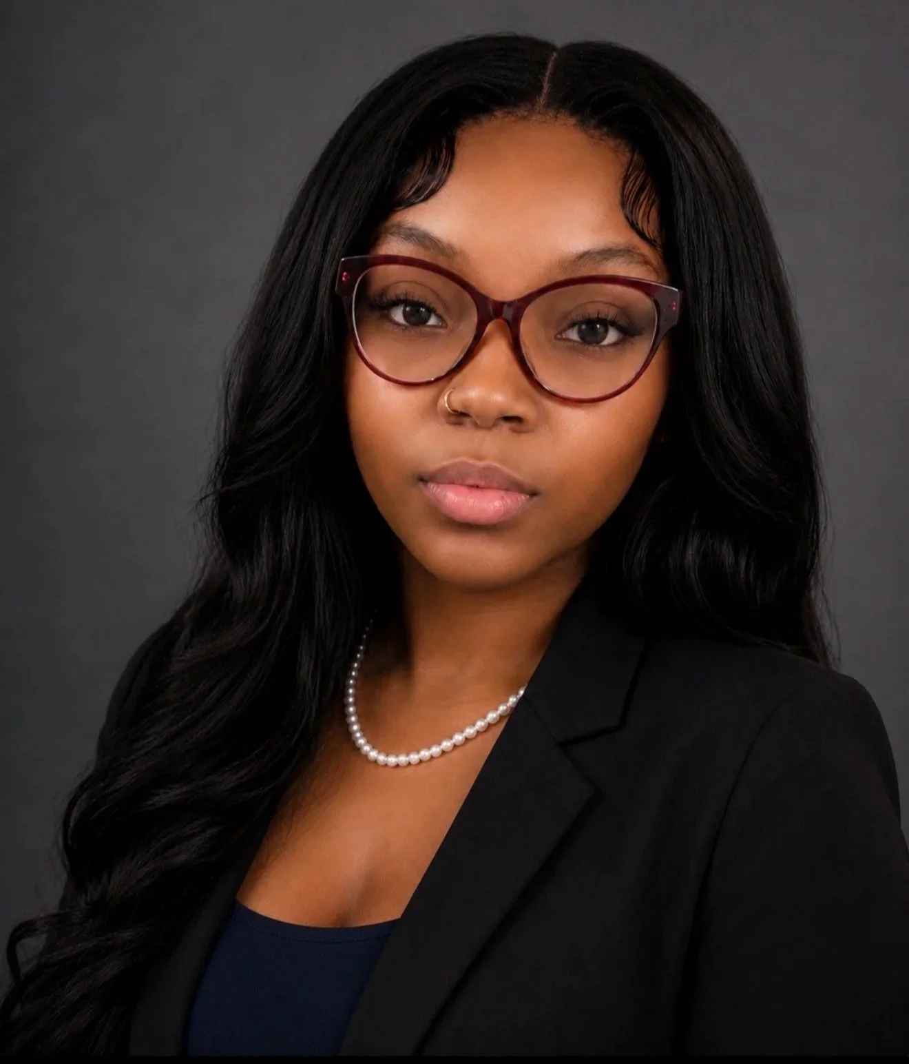 A professional portrait of a young Black woman with long wavy black hair, wearing glasses, a pearl necklace, and formal business attire against a dark background.