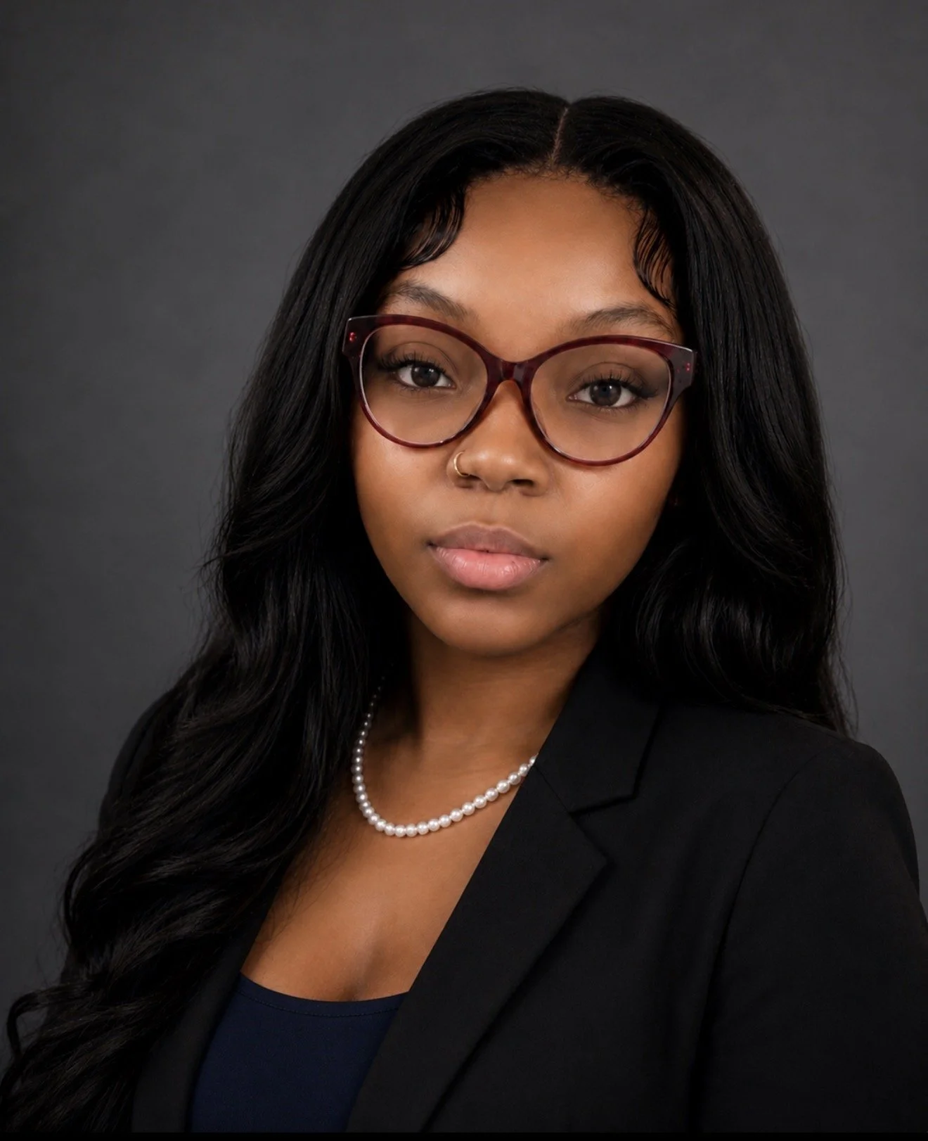 Portrait of a woman with long black hair wearing glasses, a black blazer, a pearl necklace, and a nose ring, against a dark gray background.
