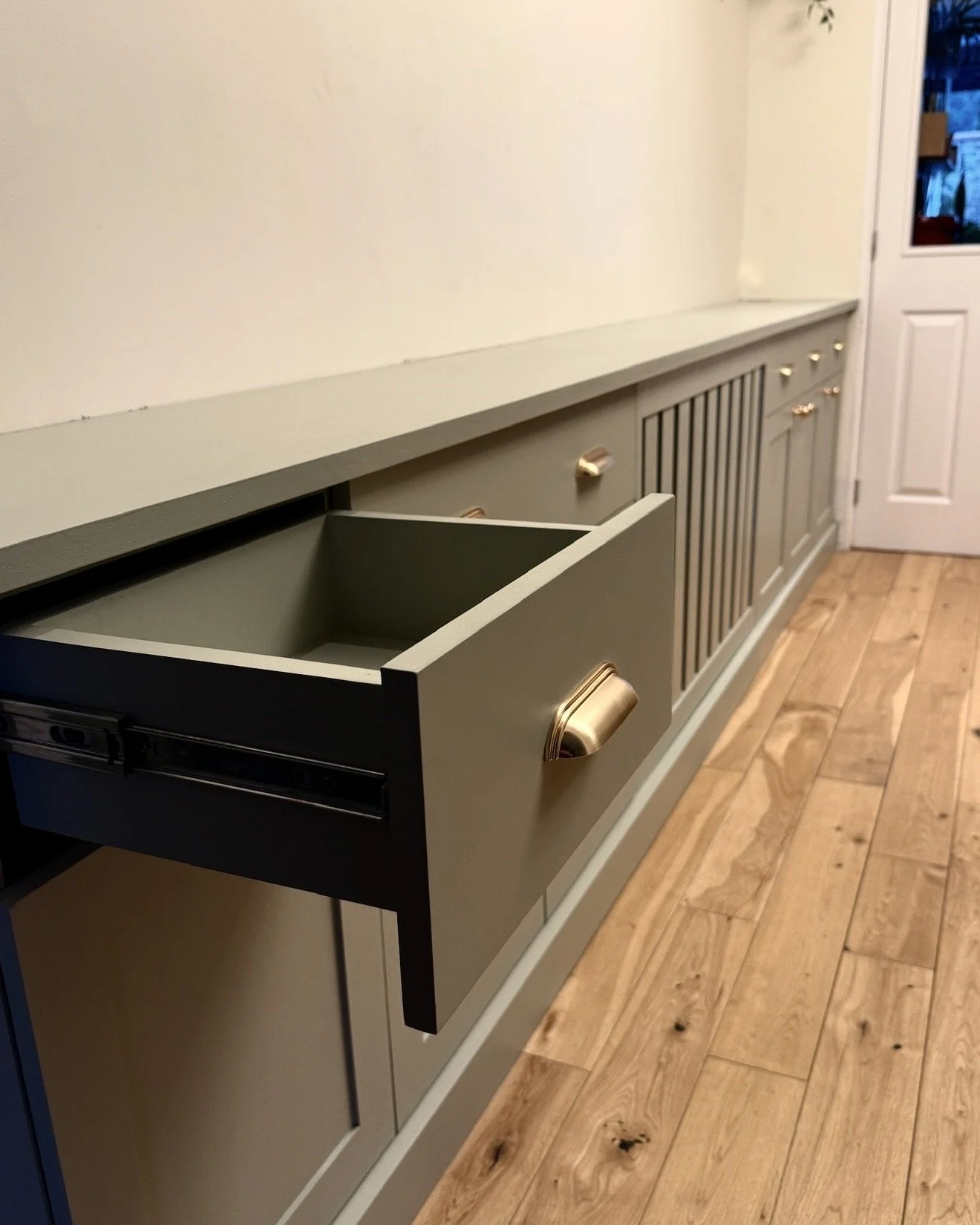 A green cabinet with brushed brass handles in a room with wooden flooring and white walls.
