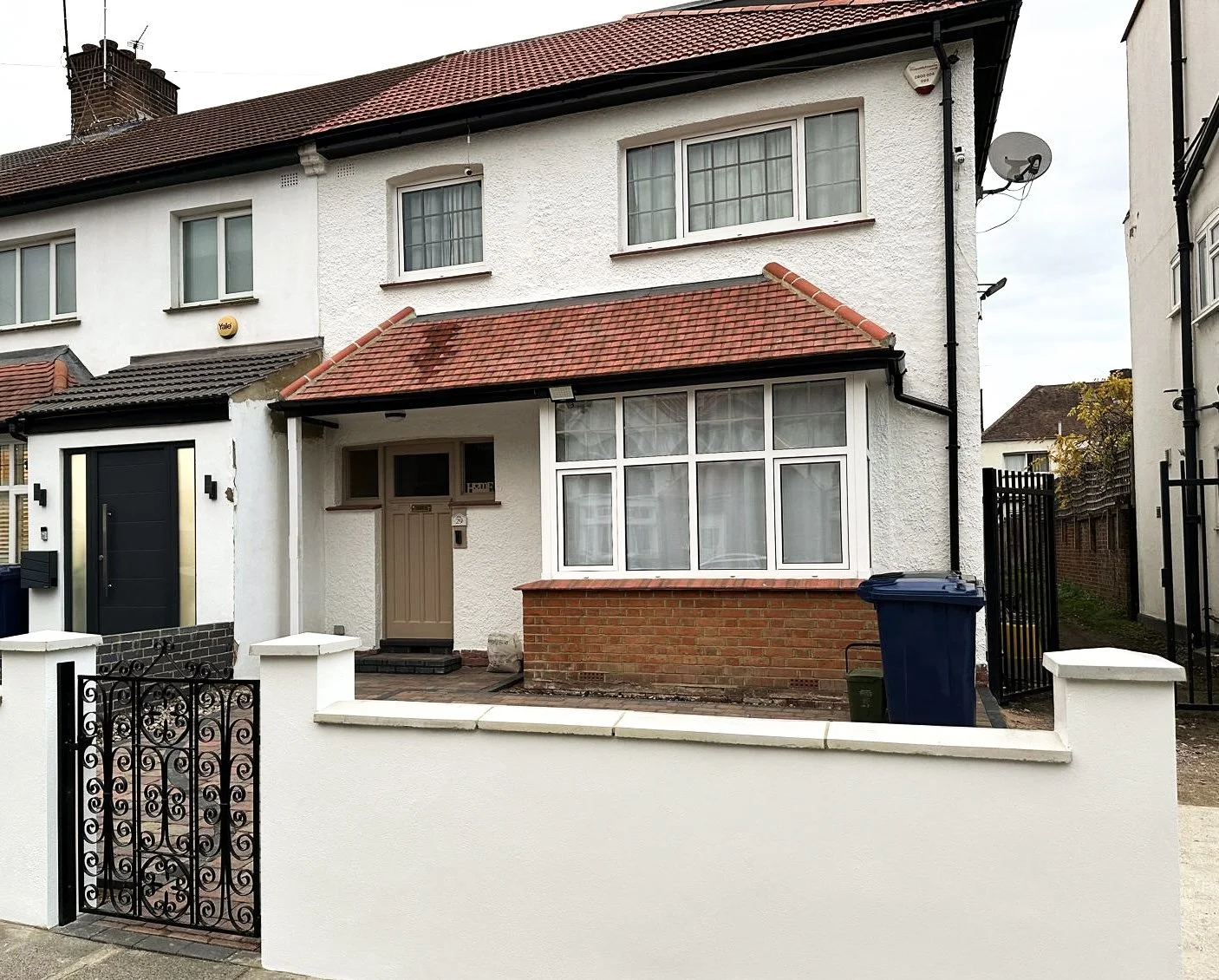 A white two-story house with red tiled roof, front door, large front bay window, and a small front yard enclosed by a white wall and black iron gate.