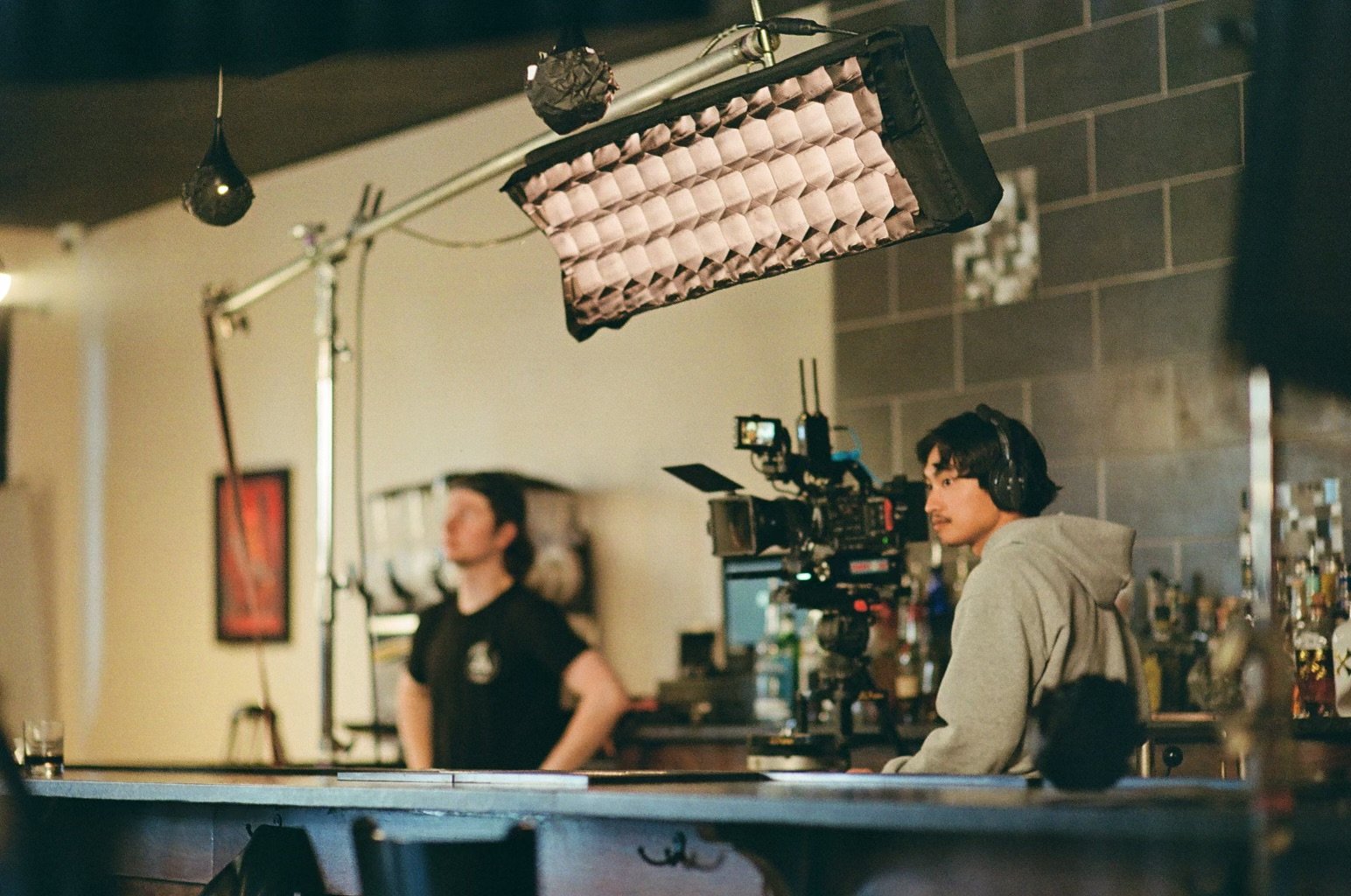 A film set with a camera operator in the foreground, wearing headphones, focusing on a scene featuring a woman in black standing in the background. Overhead lighting with a softbox is used, and the setting appears to be a bar or casual indoor space with bottles and decor on the shelves.