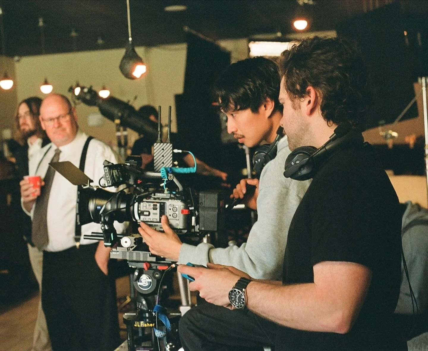 Filmmakers operating a professional video camera on set, with crew members and a man holding a red cup in the background.
