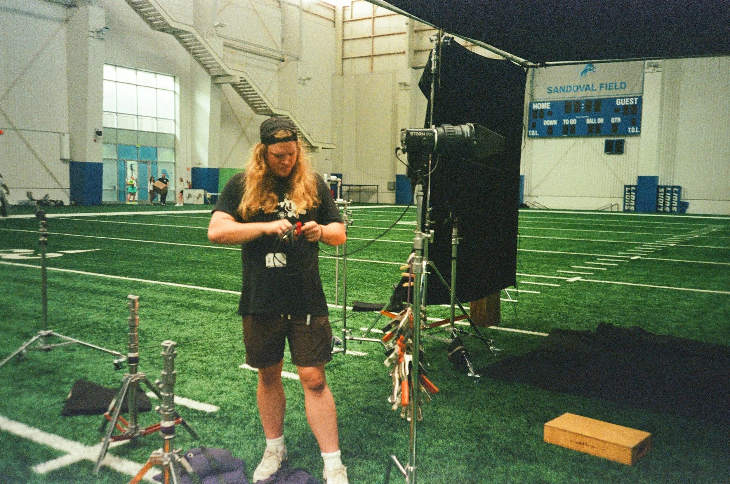A man with long blond hair, wearing a black T-shirt and shorts, is setting up film or photography equipment inside an indoor sports facility. The background shows a scoreboard reading 'Sandoval Field' and a football field with yard lines and goalposts. Several people are visible in the distance near the field's entrance.