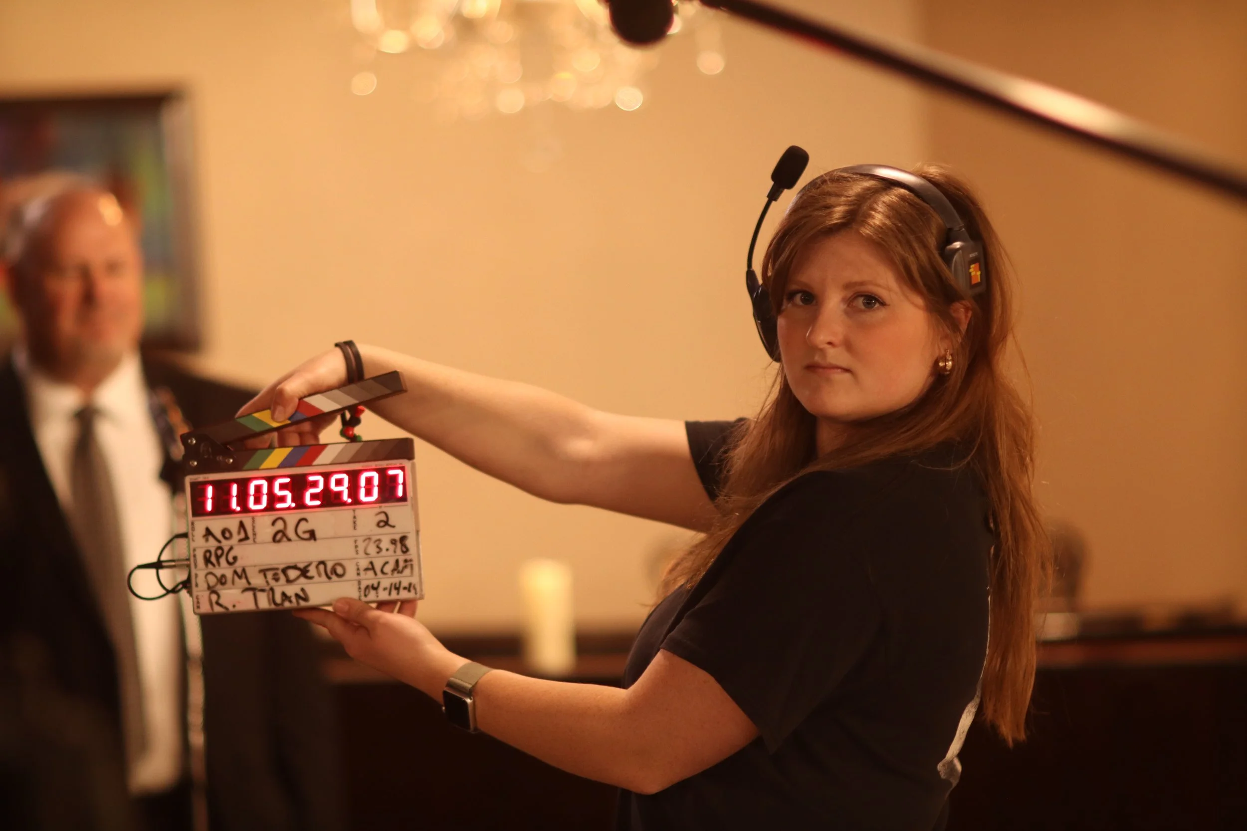 A woman with long red hair wearing a headset holds a clapperboard on a film set. She is looking slightly to the camera, and a man in a suit is blurred in the background. The clapperboard has various handwritten notes and a digital timestamp.