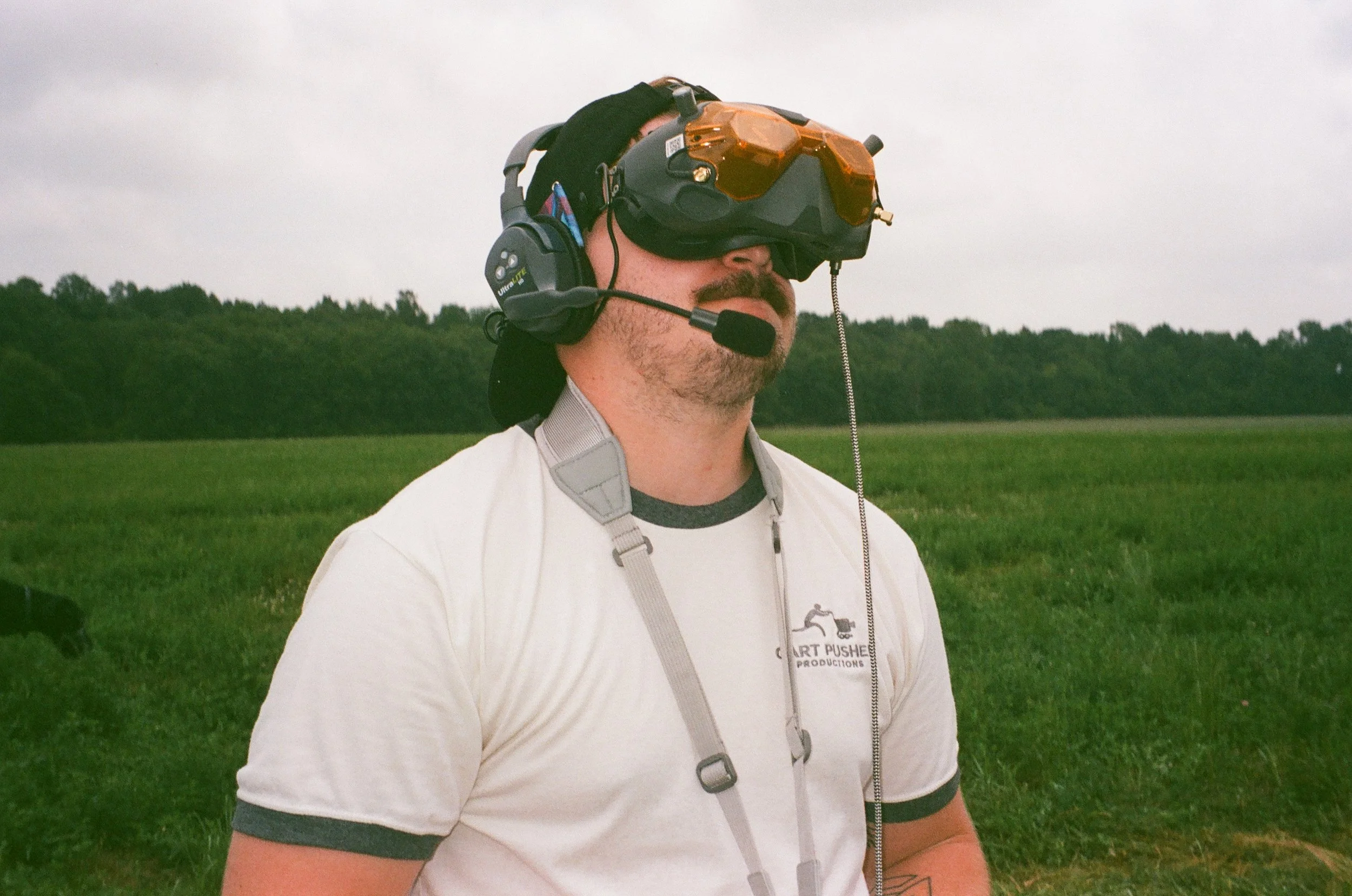 Man wearing a white T-shirt with a harness, a black helmet with orange tinted goggles, and a headset with microphone, standing in a grassy field with trees in the background.