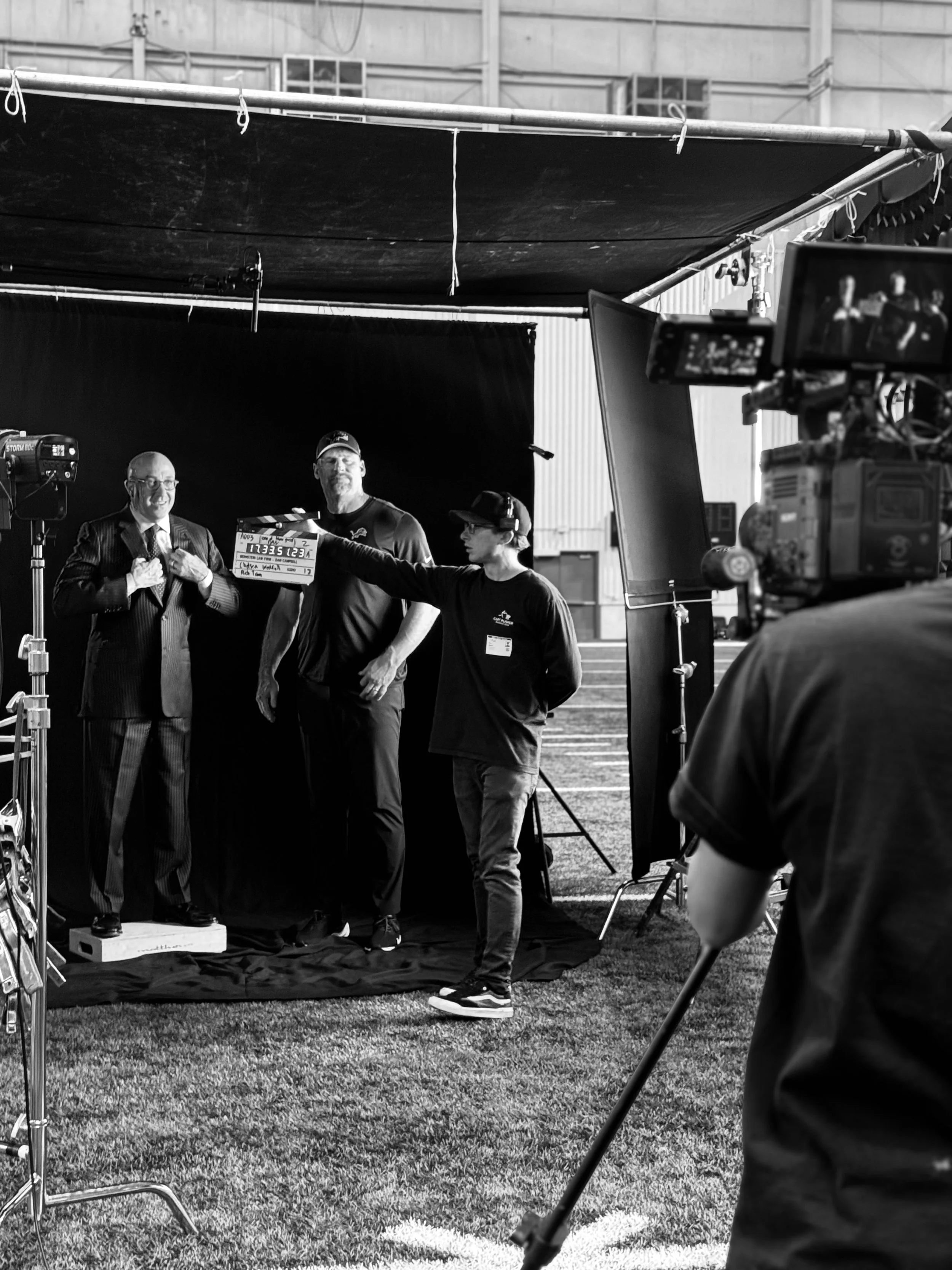 Black and white photo of a filming set with three men standing in front of a black backdrop. One man is in a suit, another in a sports shirt and cap, and the third in a t-shirt and cap. A person with a camera is in the foreground, and there is film equipment, lights, and a large black sheet hanging overhead.