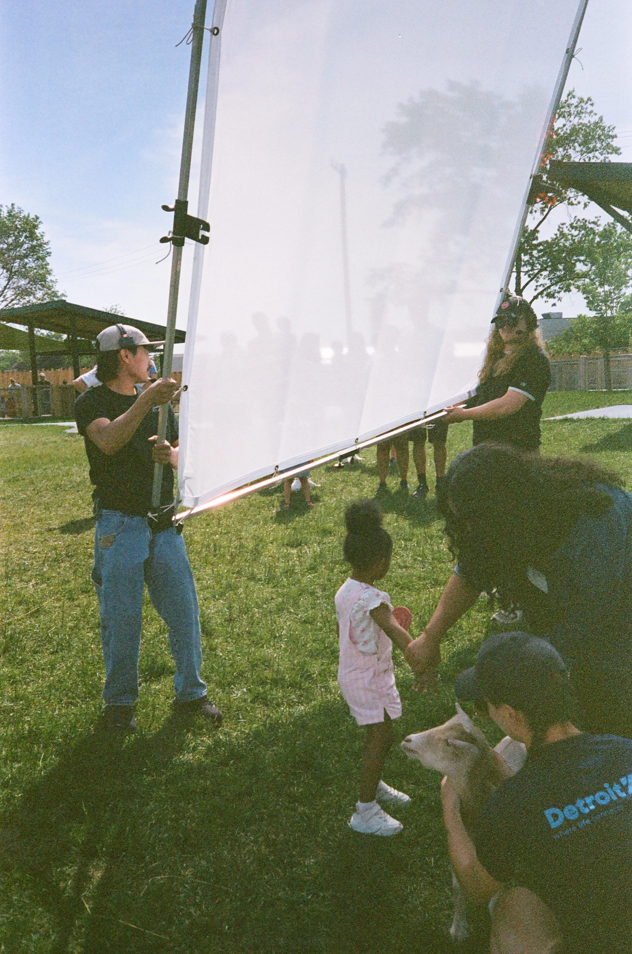 Children and adults gathering outdoors on a grassy field during daylight, with some holding a large translucent white screen or tarp, and children holding hands.
