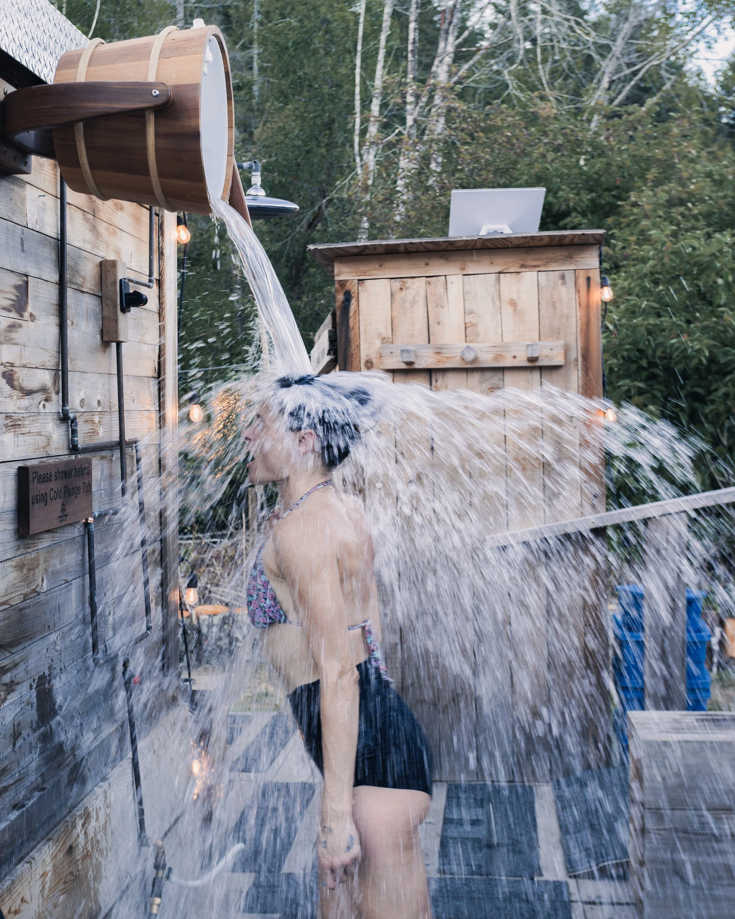 Person with short dark hair and glasses showers outdoors in a wooded area, standing under a wooden waterfall shower with water flowing over their head.