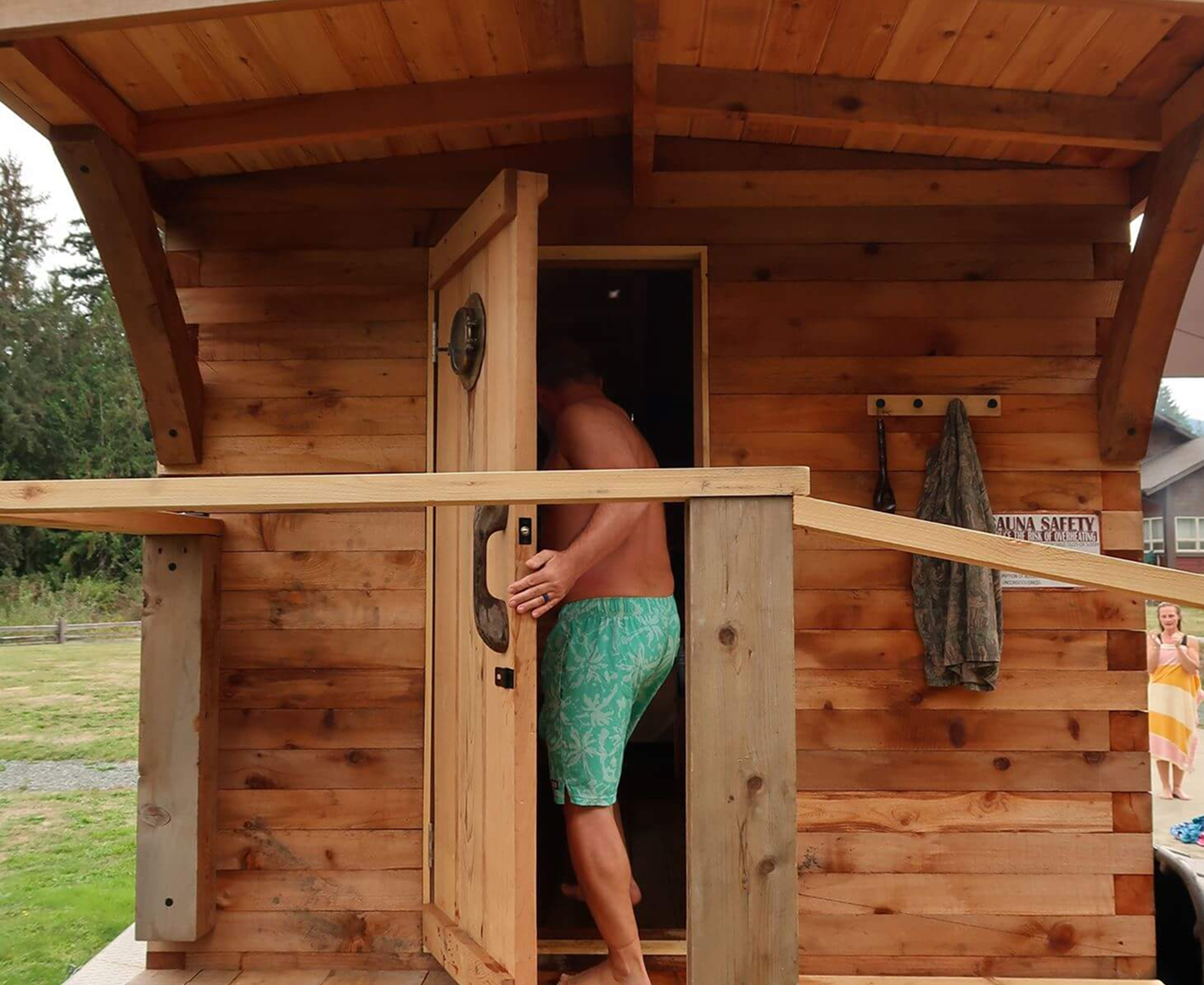 A man with a towel around his waist standing in front of a wooden sauna at an outdoor spa, with a woman in a towel on the grass nearby.