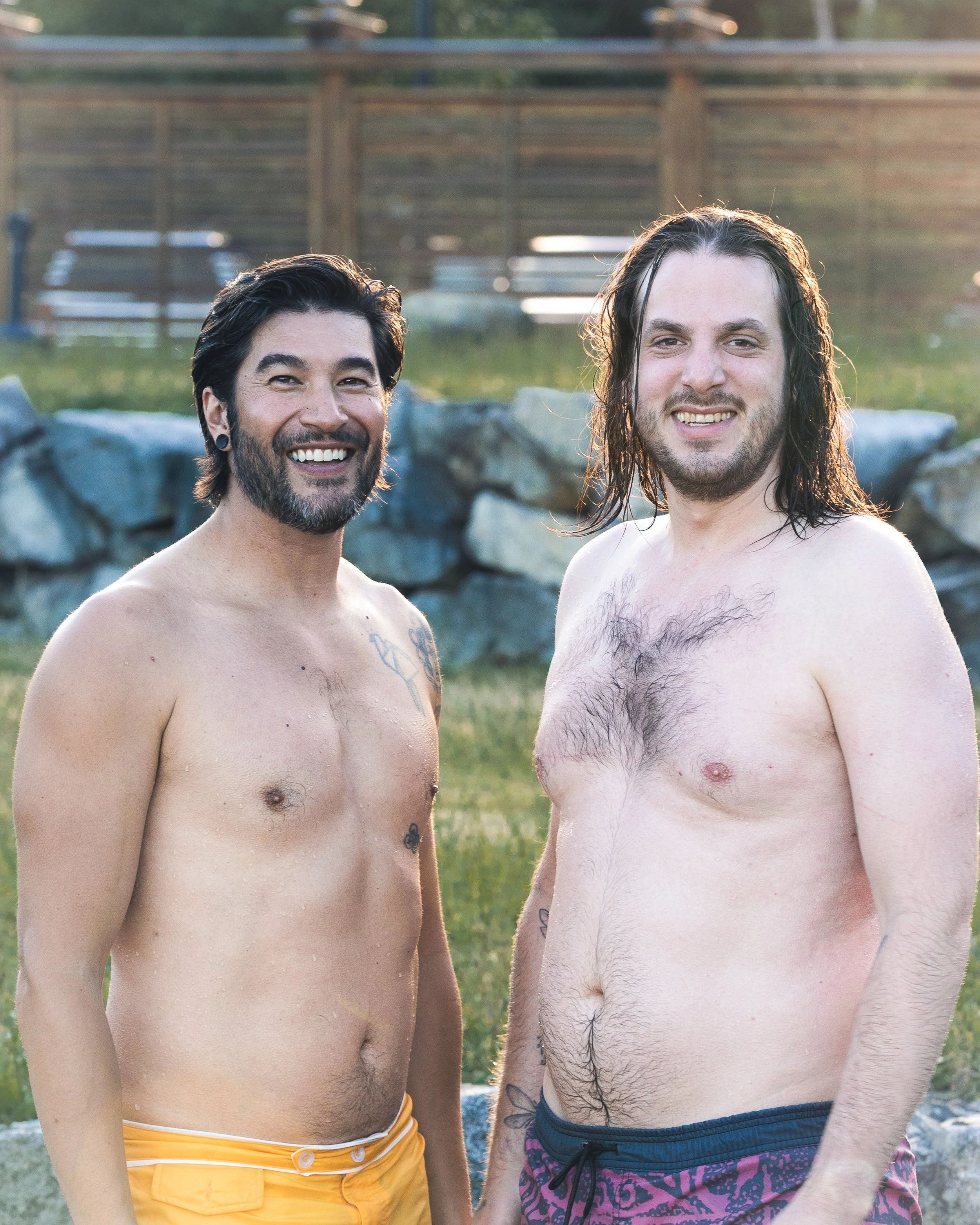 Two shirtless men smiling outdoors by a rock wall, with a wooden fence and grass in the background.