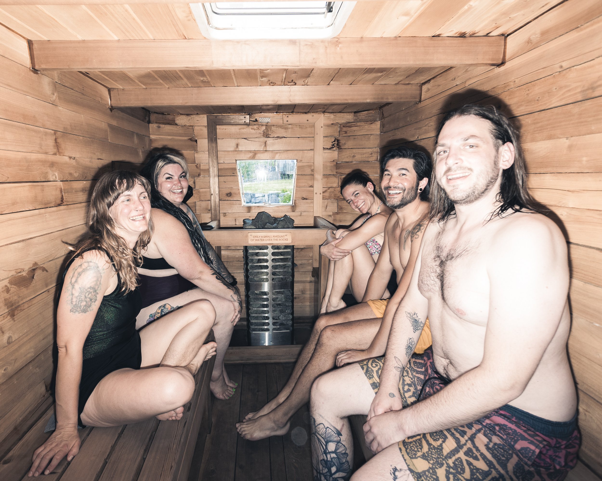 Seven people sitting in a wooden sauna, all smiling and looking at the camera, with some tattoos visible on their arms and legs.