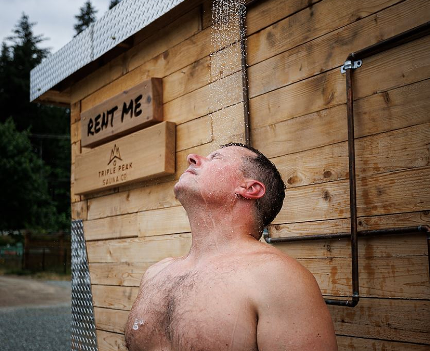 A shirtless man with short dark hair is taking an outdoor shower, standing with his head tilted back and water falling on his face, against a wood-paneled wall with signs.