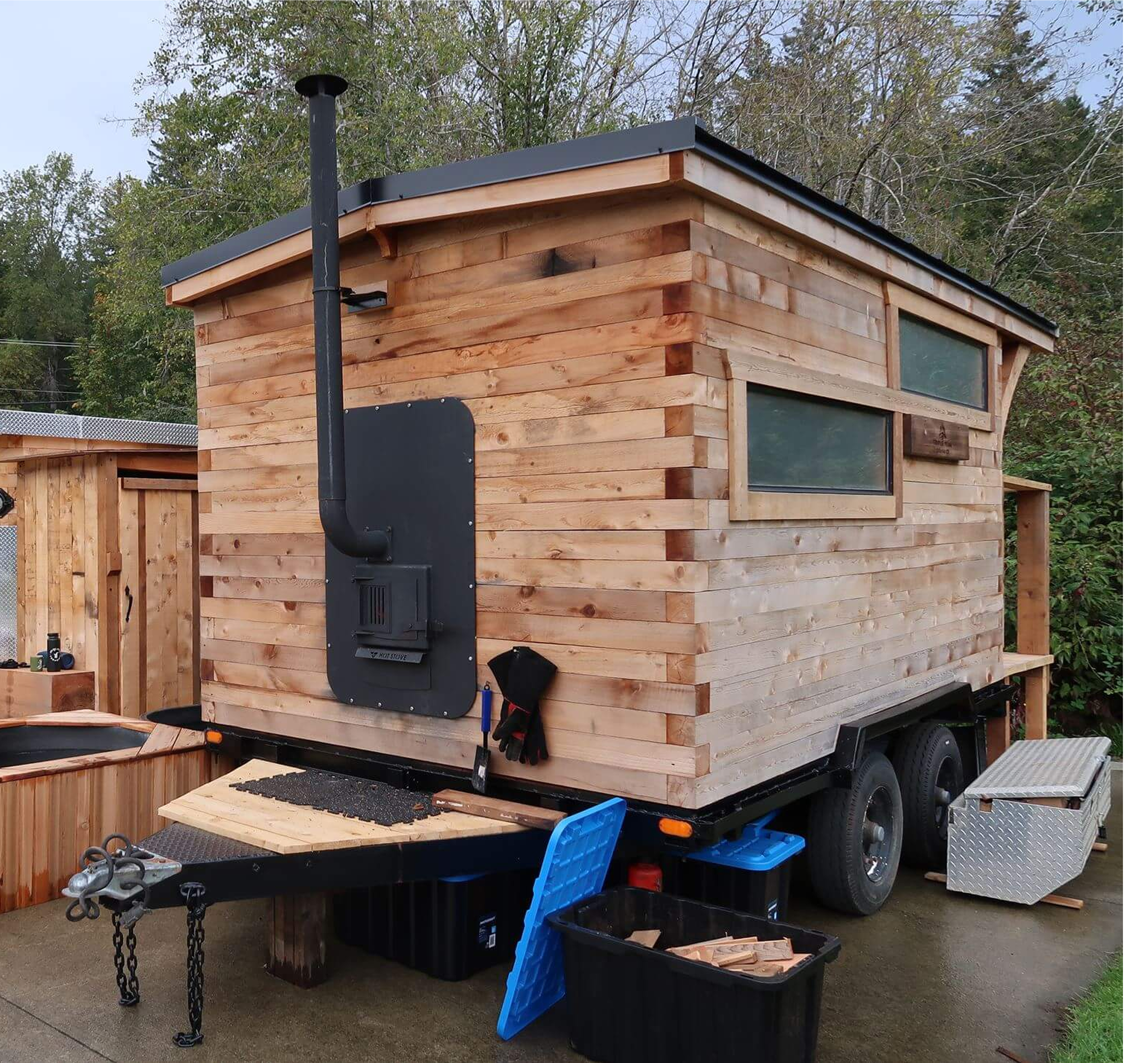 A small wooden building with a black metal roof and large windows, mounted on a trailer with a hitch and chains. There are storage bins with wood scraps and tools nearby.