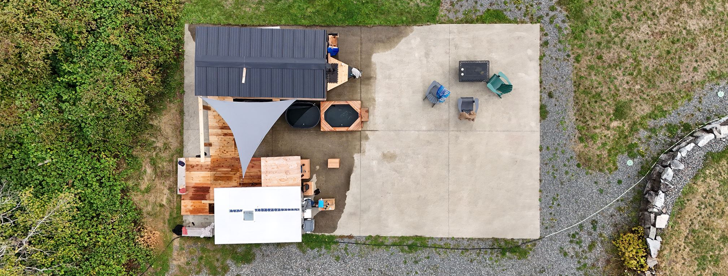 An aerial view of a backyard patio with a wooden deck, a shade sail, a white-roofed small building, and outdoor furniture including chairs and a table. Surrounding greenery and a gravel pathway are visible.
