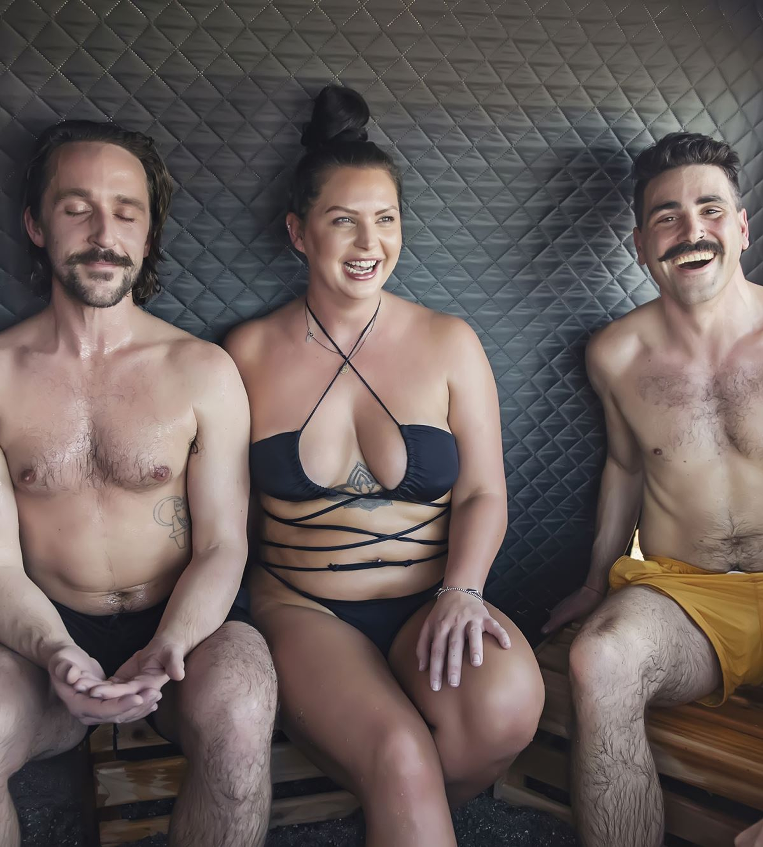Three people sitting on a bed or couch, topless, smiling and relaxing. The person in the middle is a woman with dark hair in a ponytail, and the two men on either side are shirtless, both with facial hair. The background has a black, quilted-textured surface.