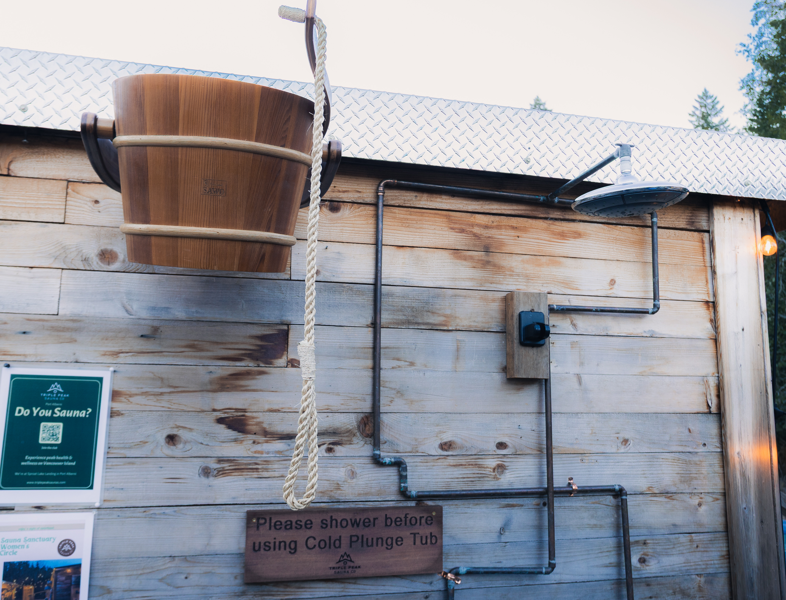 Wooden outdoor shower with a sign indicating to shower before using the cold plunge tub, set against a wooden wall with posters and a metal shower head.