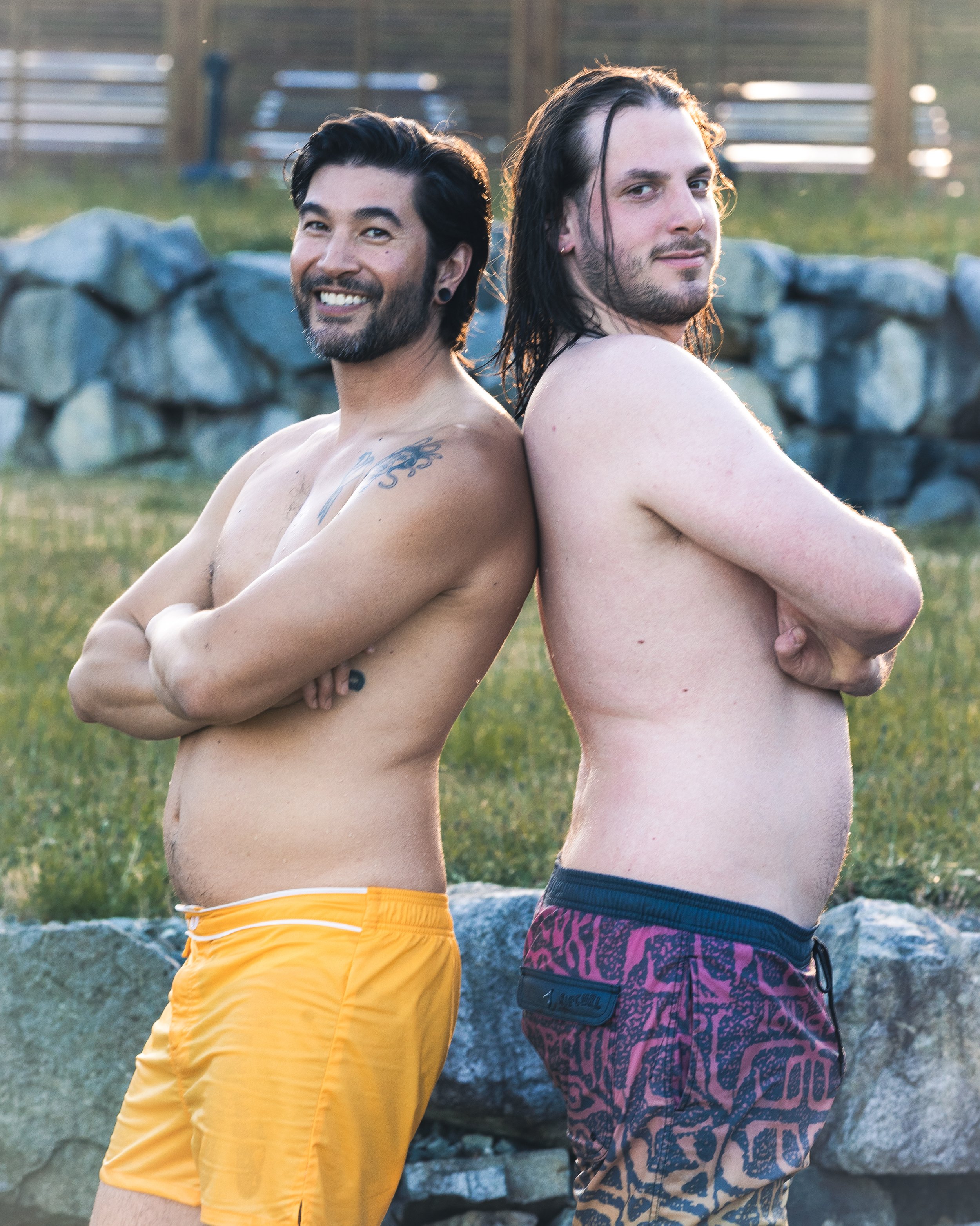 Two shirtless men standing back-to-back outdoors in front of a stone wall, smiling and looking at the camera during sunset.